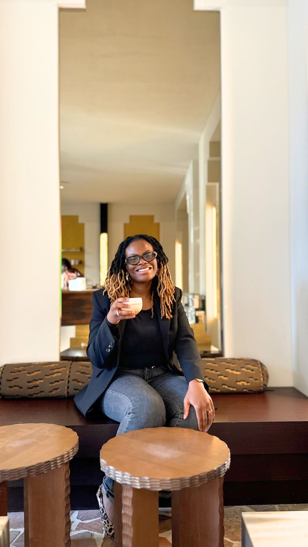 Femme entrepreneuse souriante en blazer noir, tenant une tasse de café, assise dans un salon élégant d'un hôtel.