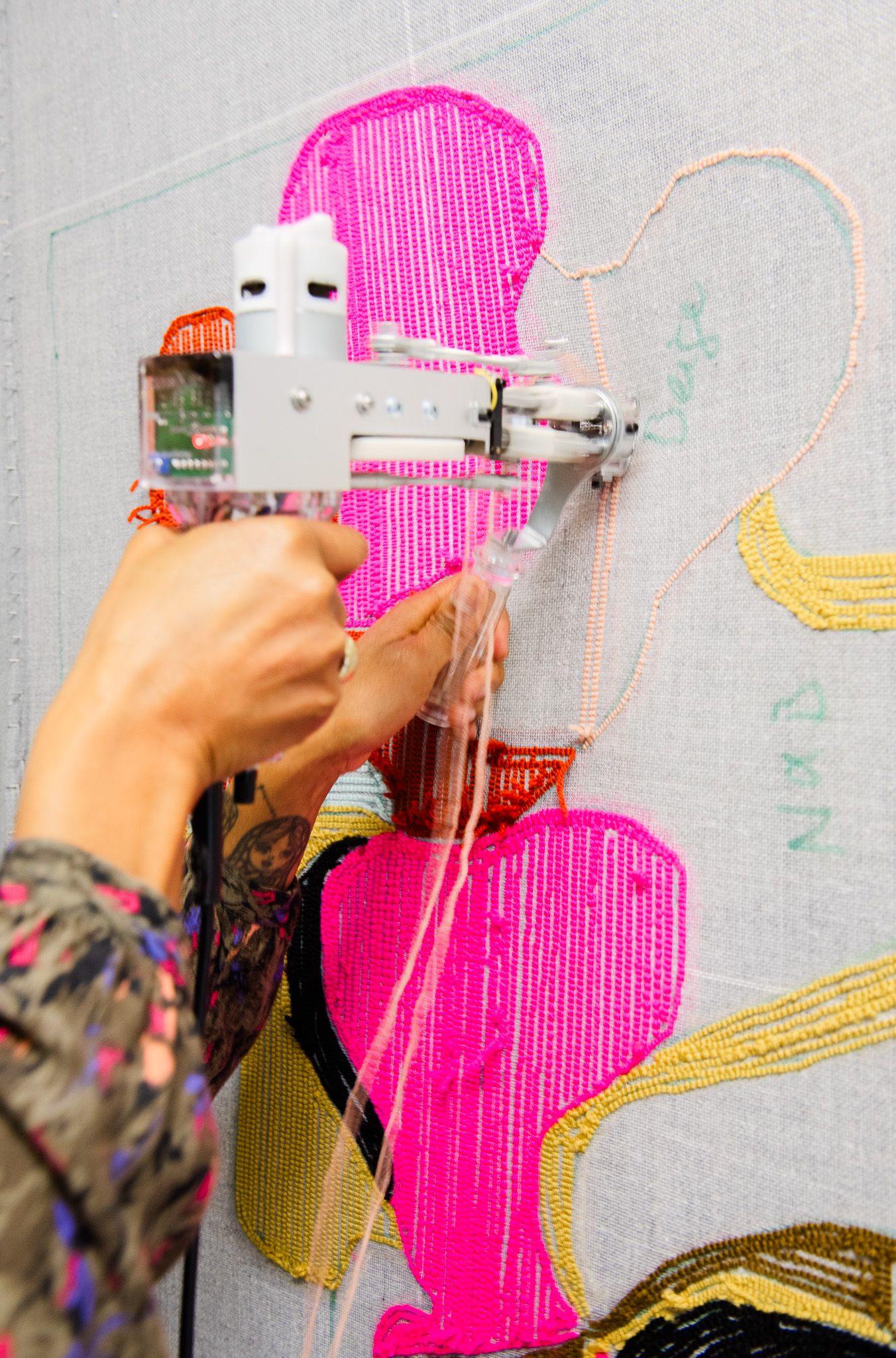 Close-up of tufting gun creating bright pink and orange abstract pattern on textile canvas artwork.