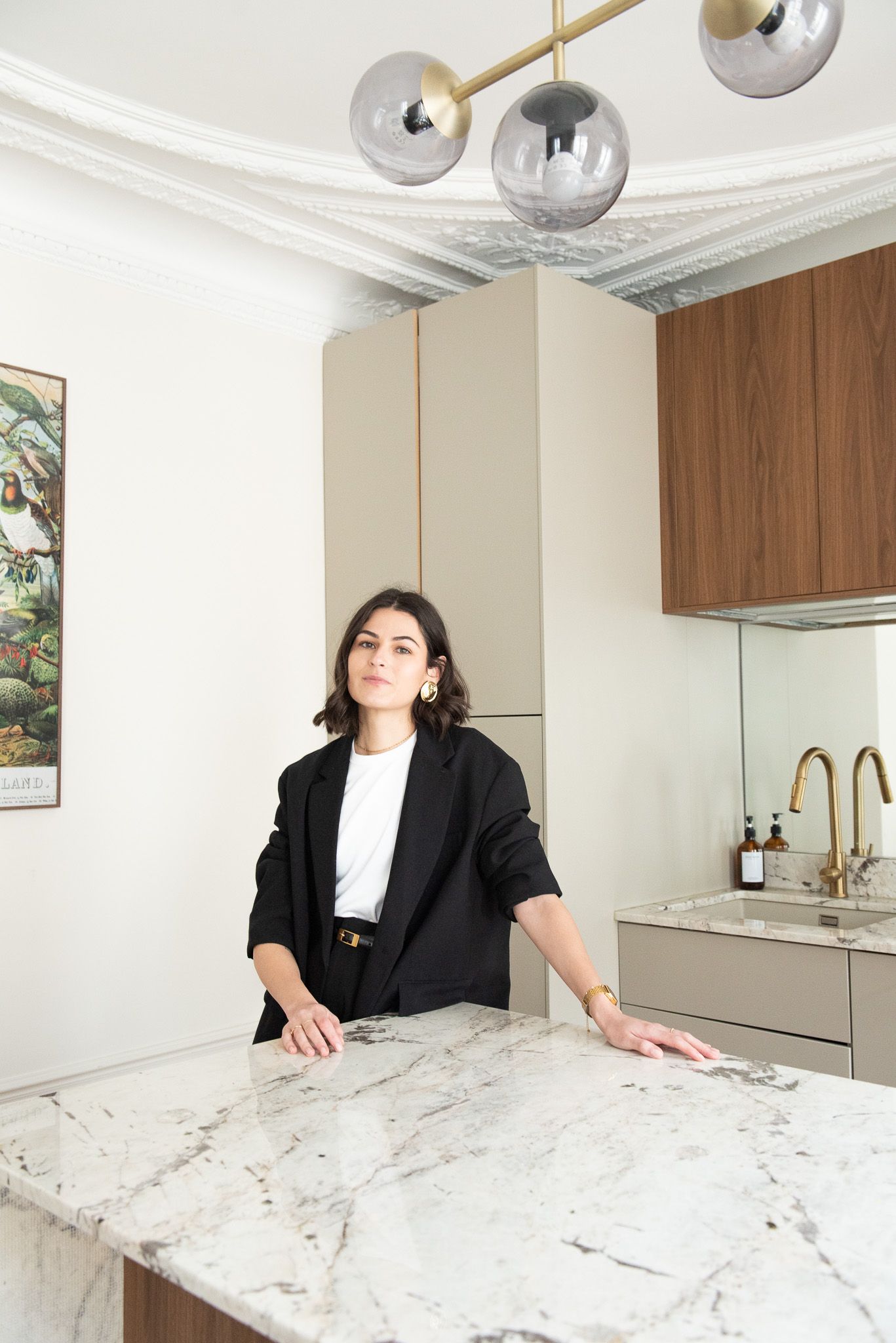 Woman in black blazer stands at marble kitchen island beneath globe pendant light in modern kitchen with brass fixtures.