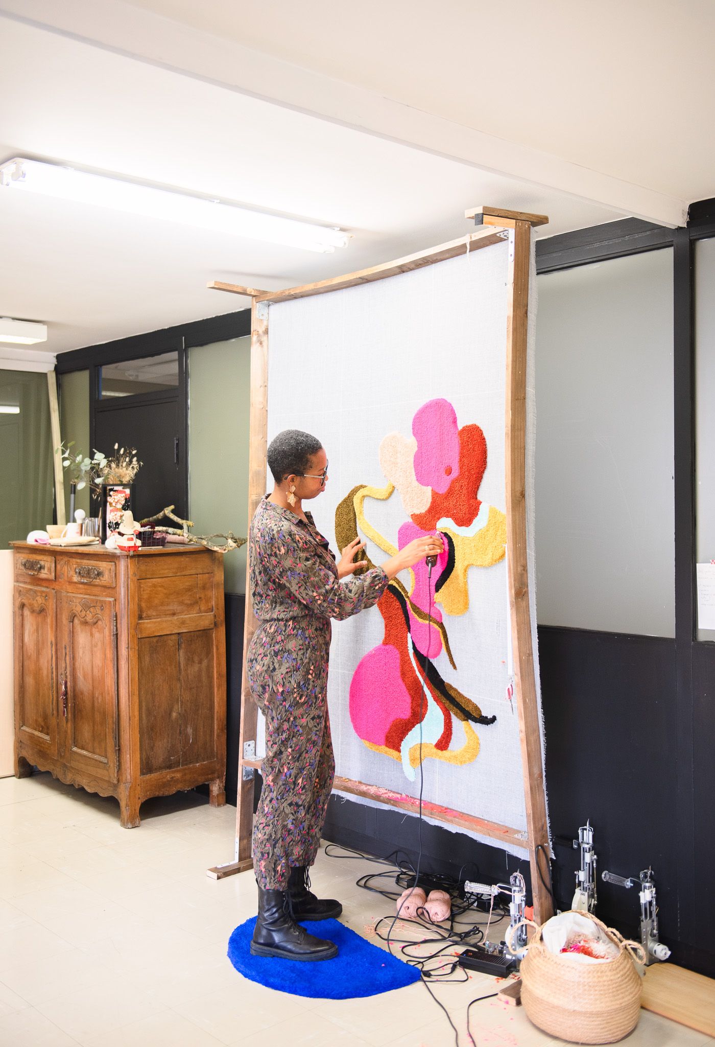Artist works on large colorful abstract tufted textile hanging on wooden frame in studio space.
