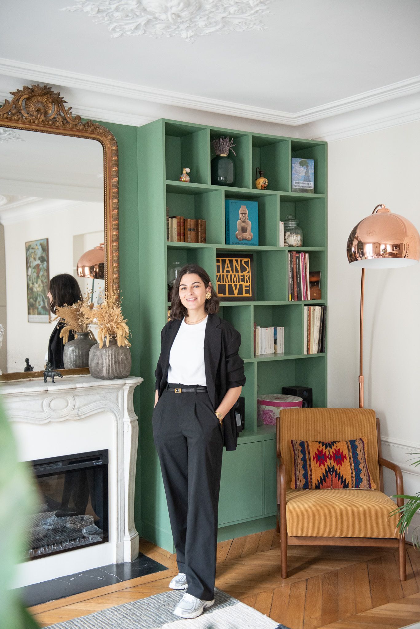 Woman in black suit standing in front of green built-in bookshelf in elegant Parisian living room with fireplace.
