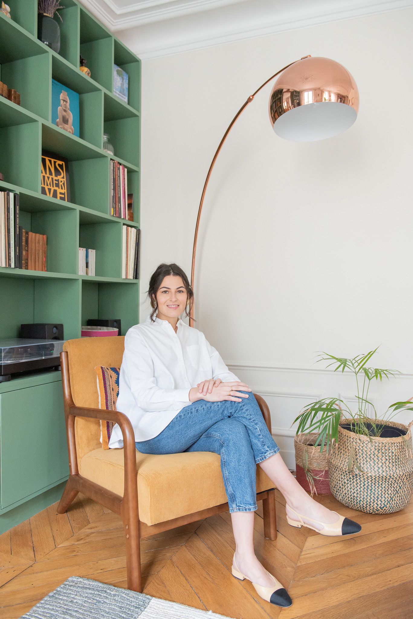 Woman sitting in a vintage armchair beside a copper arc lamp and green bookshelf in a stylish Parisian living room.