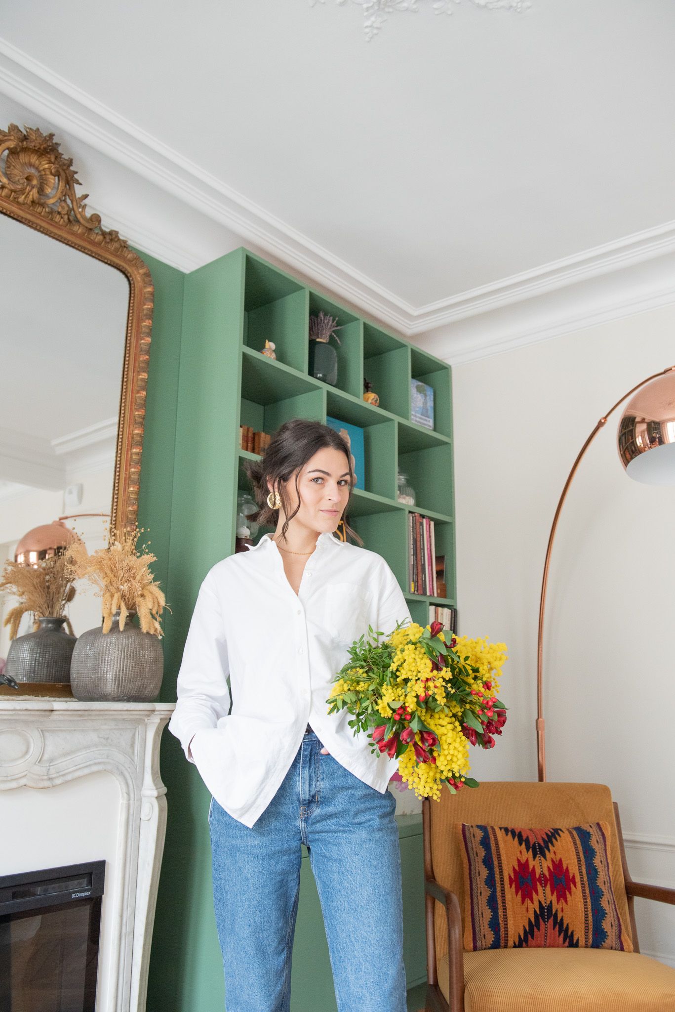 Woman in white shirt holding yellow bouquet standing near green bookshelf and ornate gold mirror.
