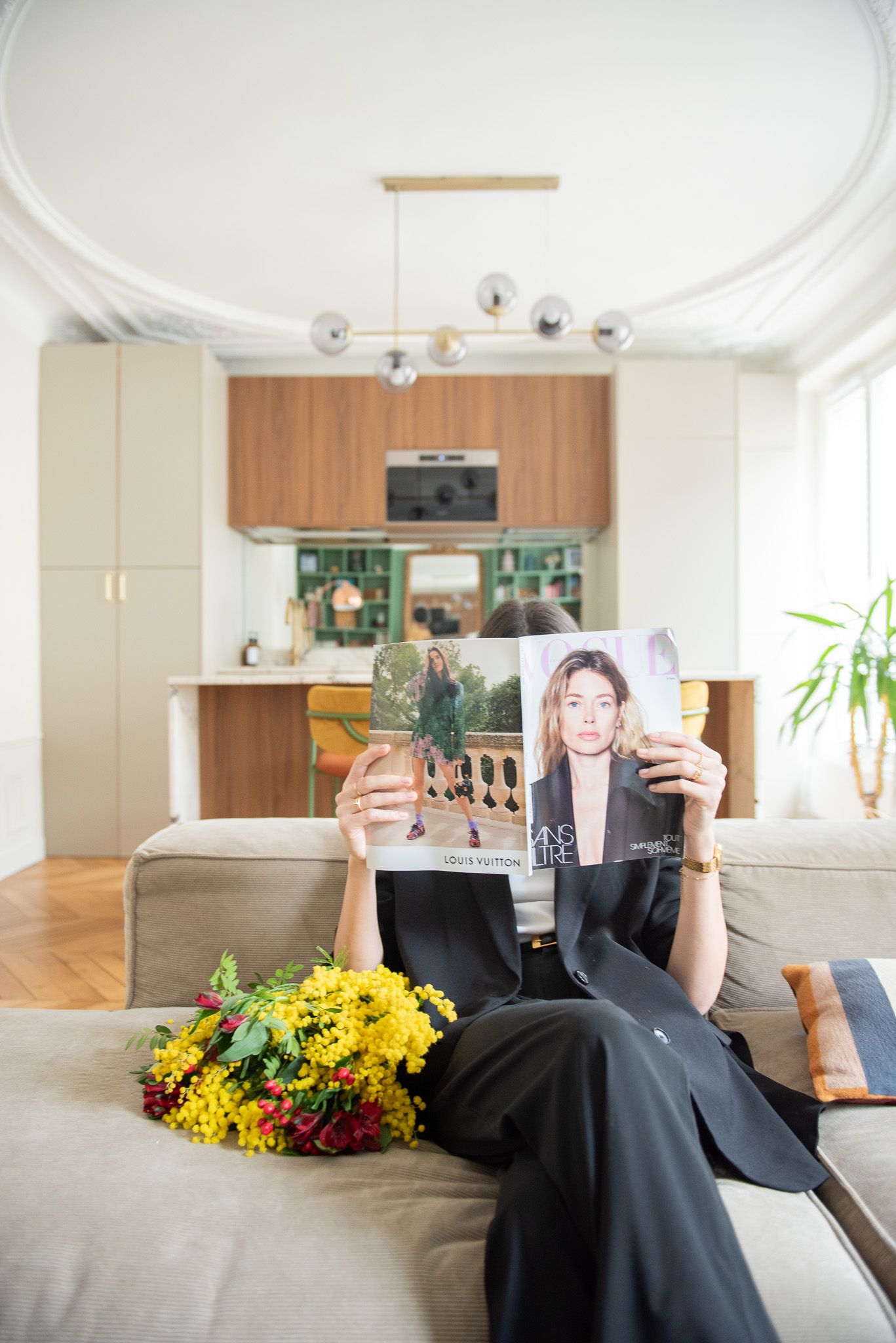 Woman in black outfit reading a fashion magazine on a beige sofa in a modern open-plan living room with yellow flowers.