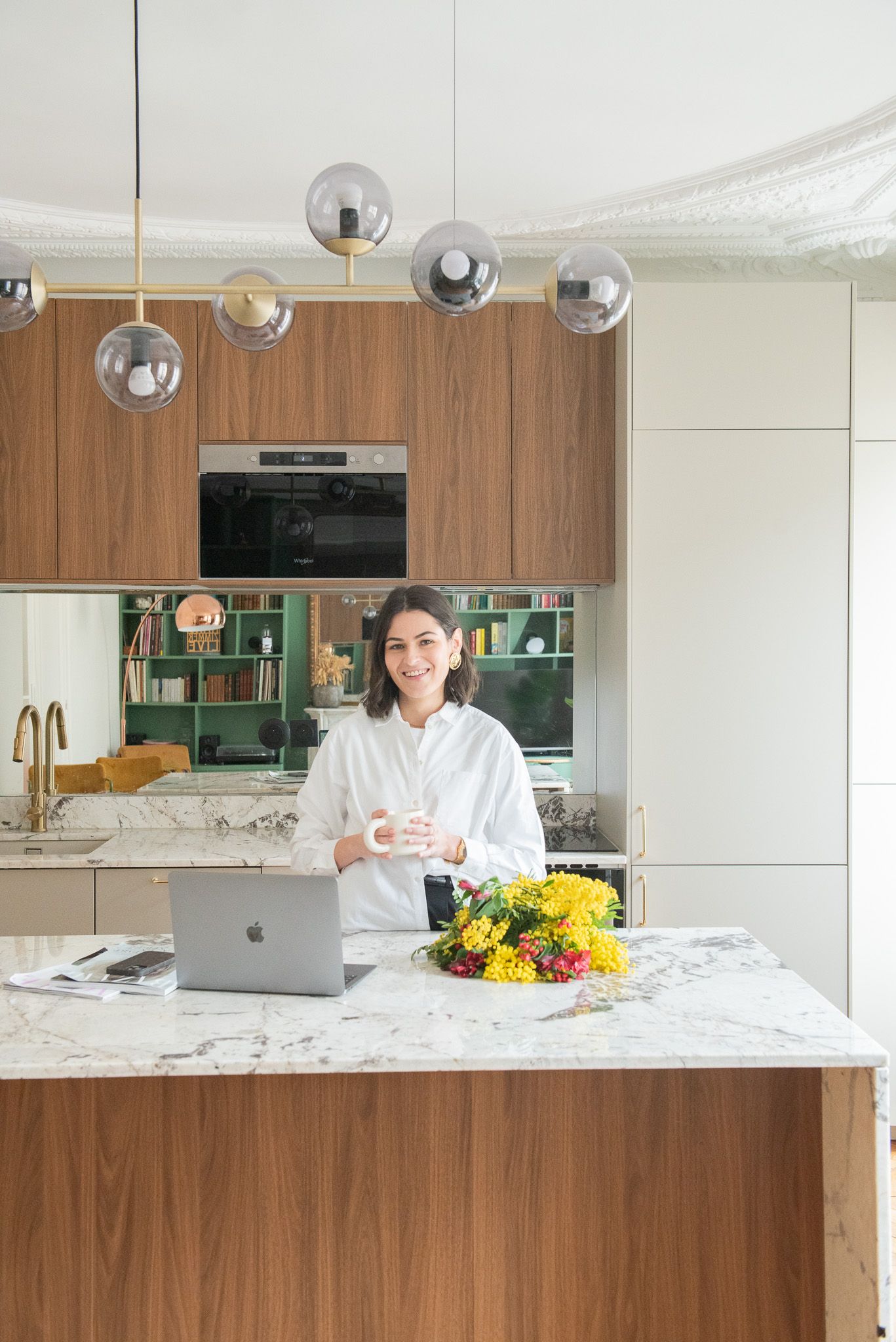 Woman smiling at marble kitchen island with laptop and flowers, modern wood cabinets and globe pendant lights.