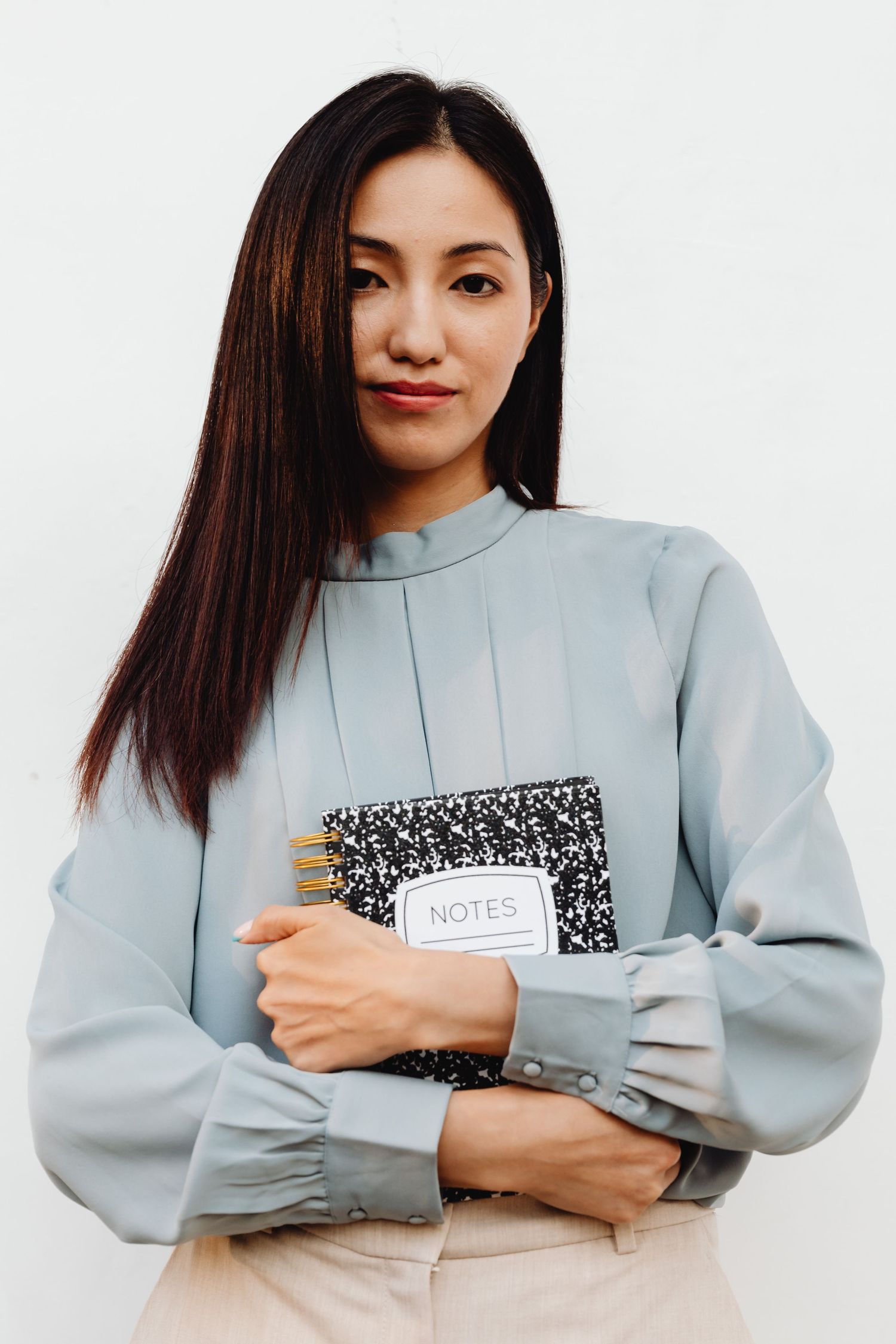Asian woman in light blue blouse holding black Notes composition notebook against white wall.