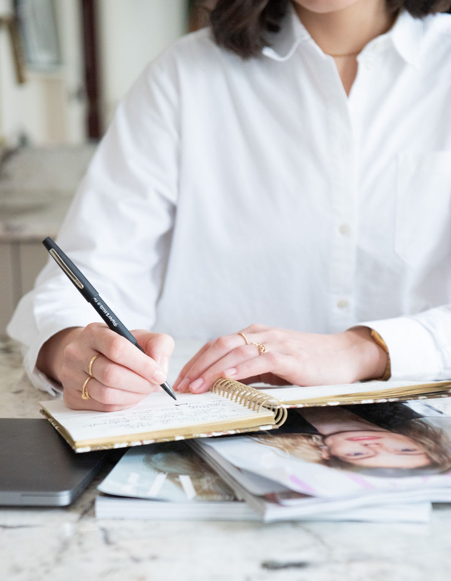 Woman in white shirt writing in spiral notebook on marble desk with rings on fingers.