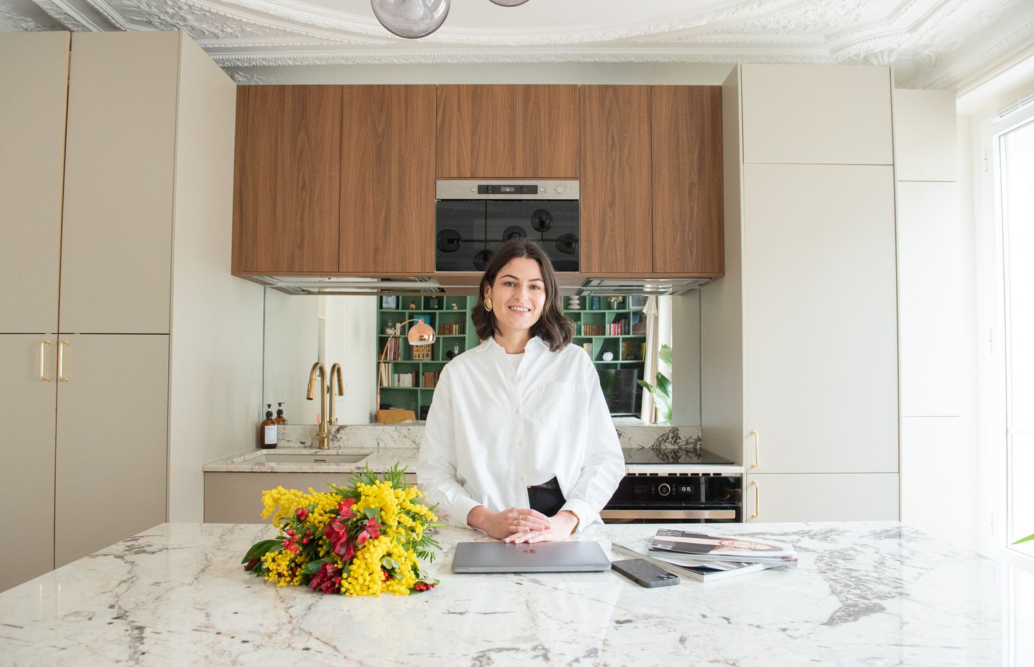 Woman smiling in modern kitchen with marble island, wood cabinets, and yellow flowers.