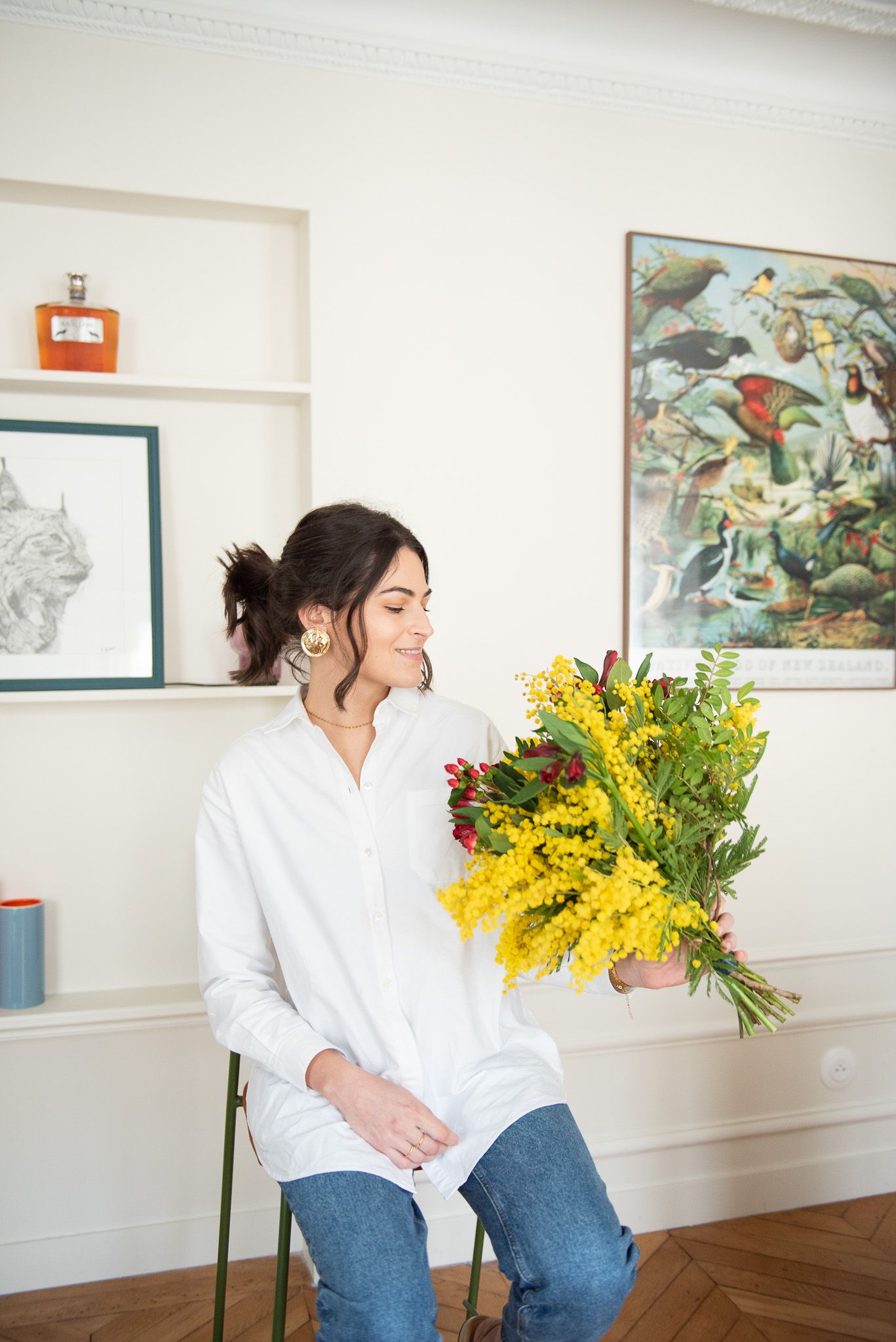 Woman in white shirt smiling while holding vibrant yellow mimosa flower bouquet indoors.