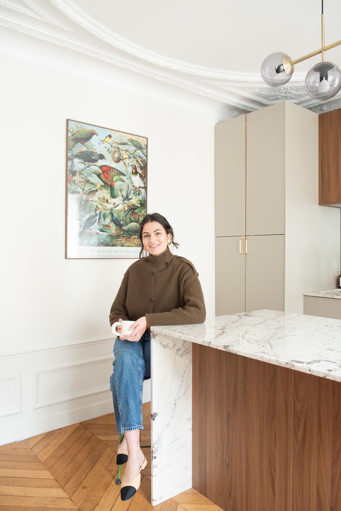 Woman in brown sweater and jeans leaning on marble kitchen island beneath modern pendant light.