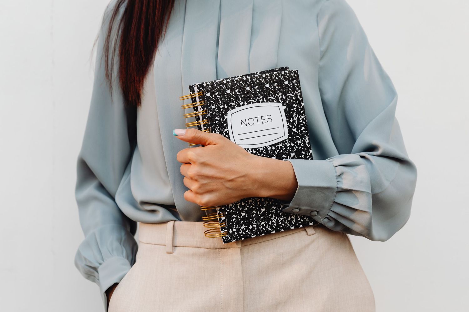 Woman in light blue blouse holding black composition notebook and gold pens against white wall.