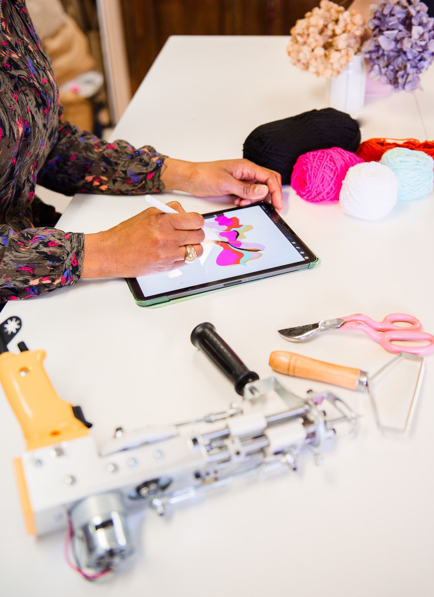Artist designs colorful textile patterns on tablet surrounded by yarn balls and tufting tools on white table.
