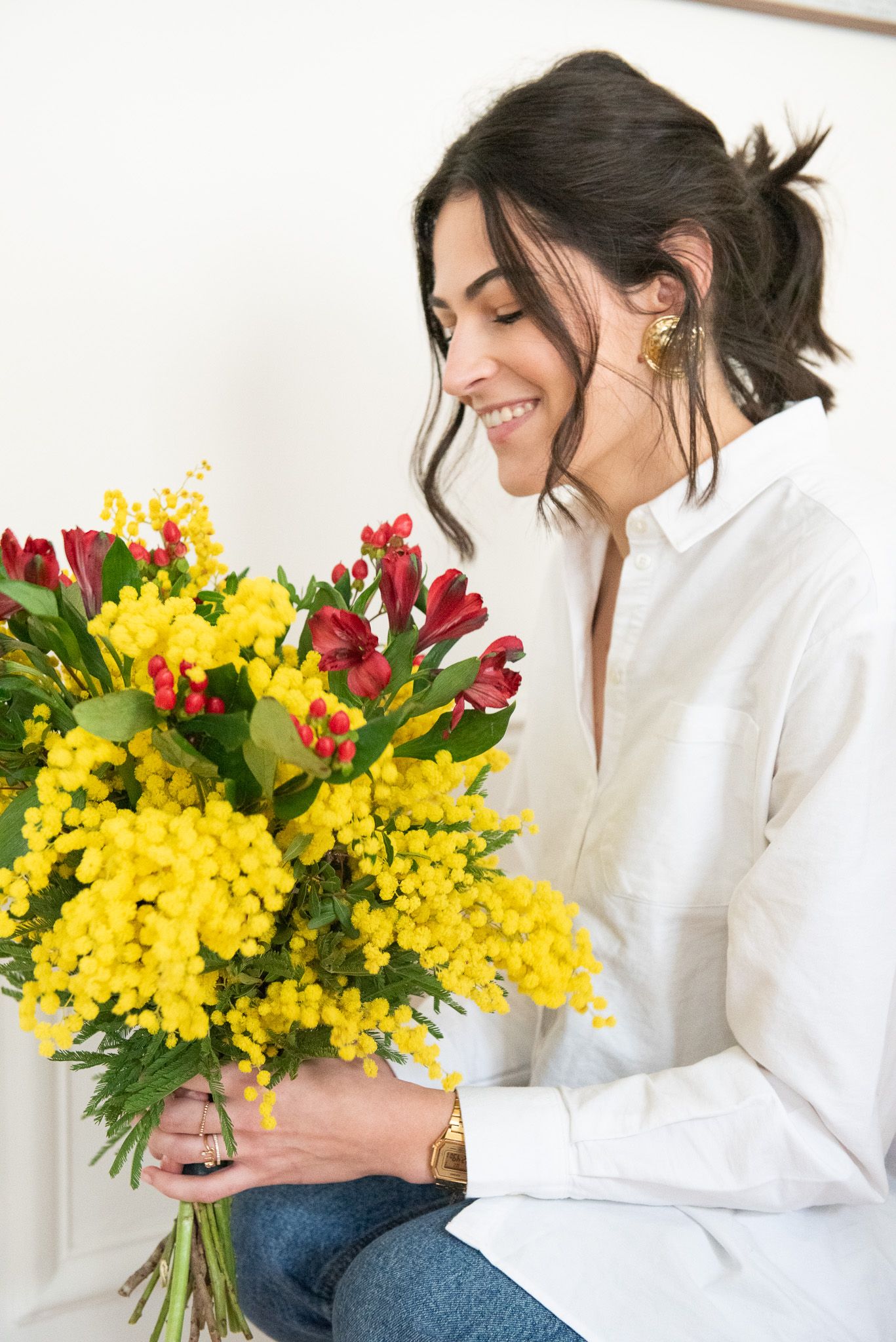 Smiling woman holding a vibrant bouquet of yellow mimosa and red alstroemeria flowers, wearing a white shirt and gold jewelry.