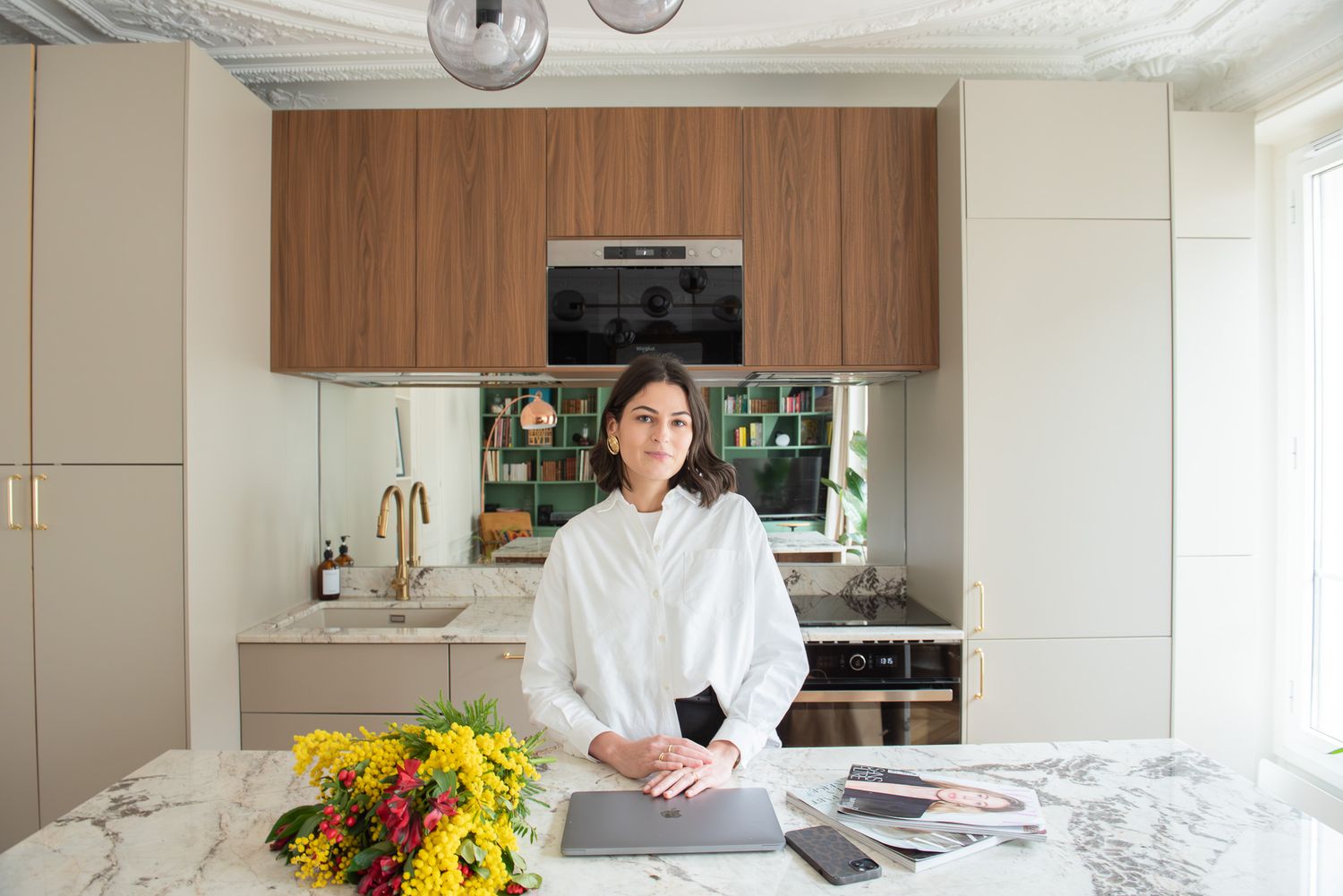 Woman in white shirt standing at marble kitchen island with yellow flowers and laptop.