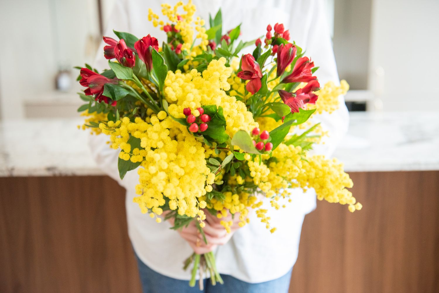 Hands holding vibrant bouquet of yellow mimosa and red tulips in bright kitchen setting.