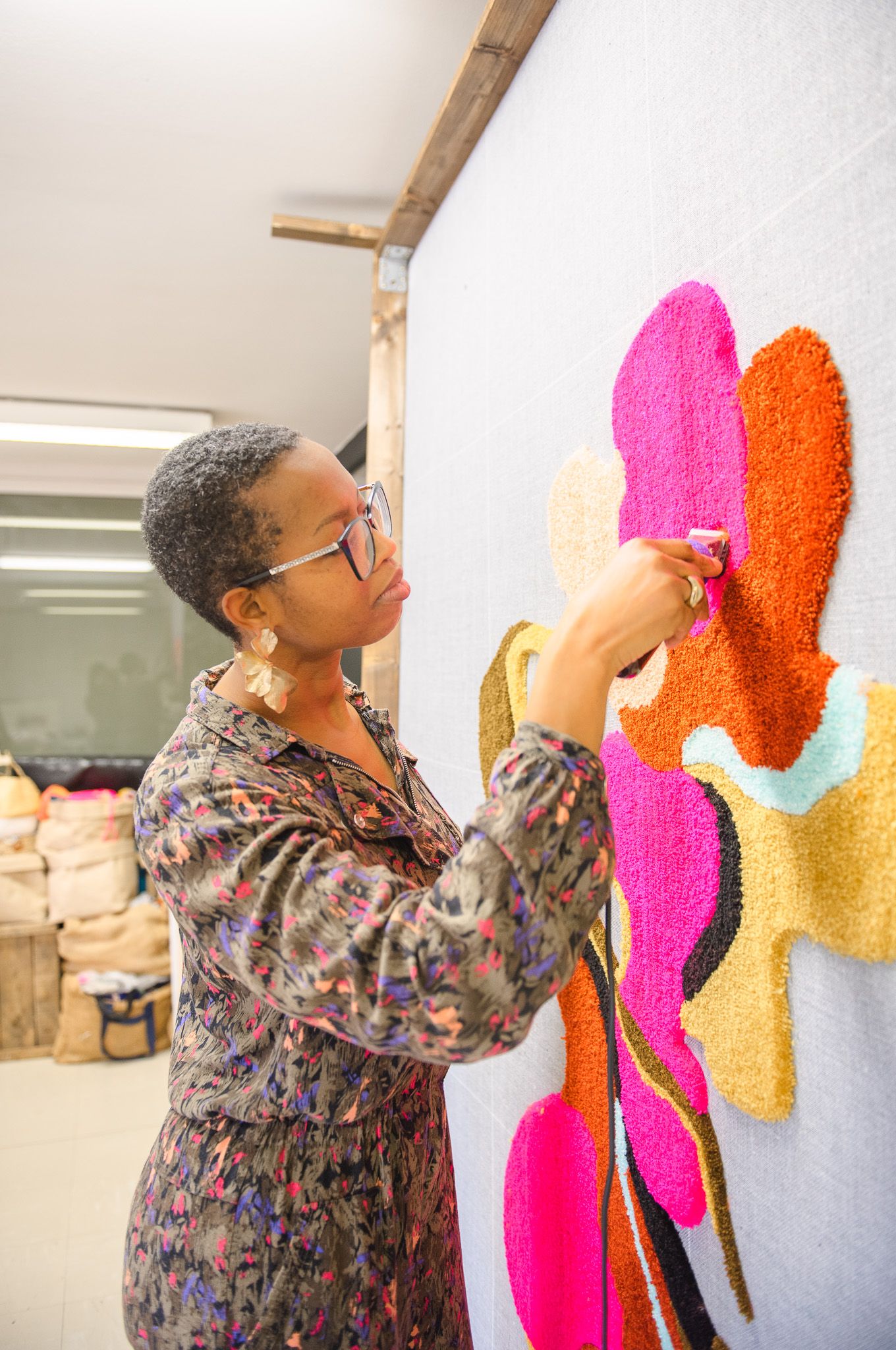 A woman artist carefully works on a vibrant colorful textile wall art piece featuring bold floral shapes in pink, orange, and teal.
