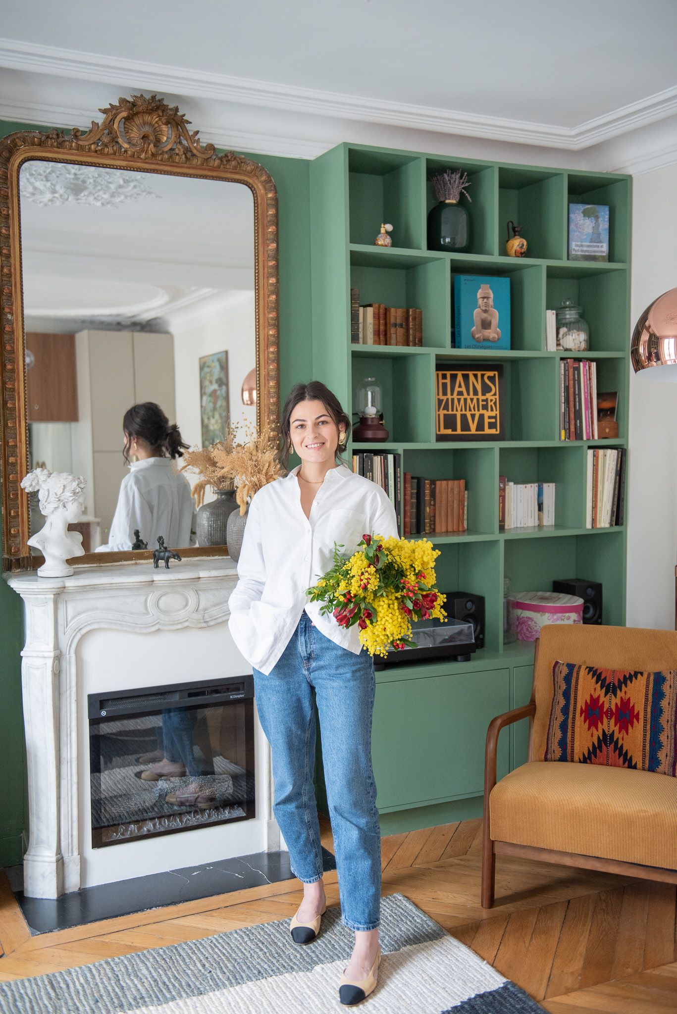 Woman holding yellow flowers in a stylish living room with green built-in shelves and ornate gold mirror.