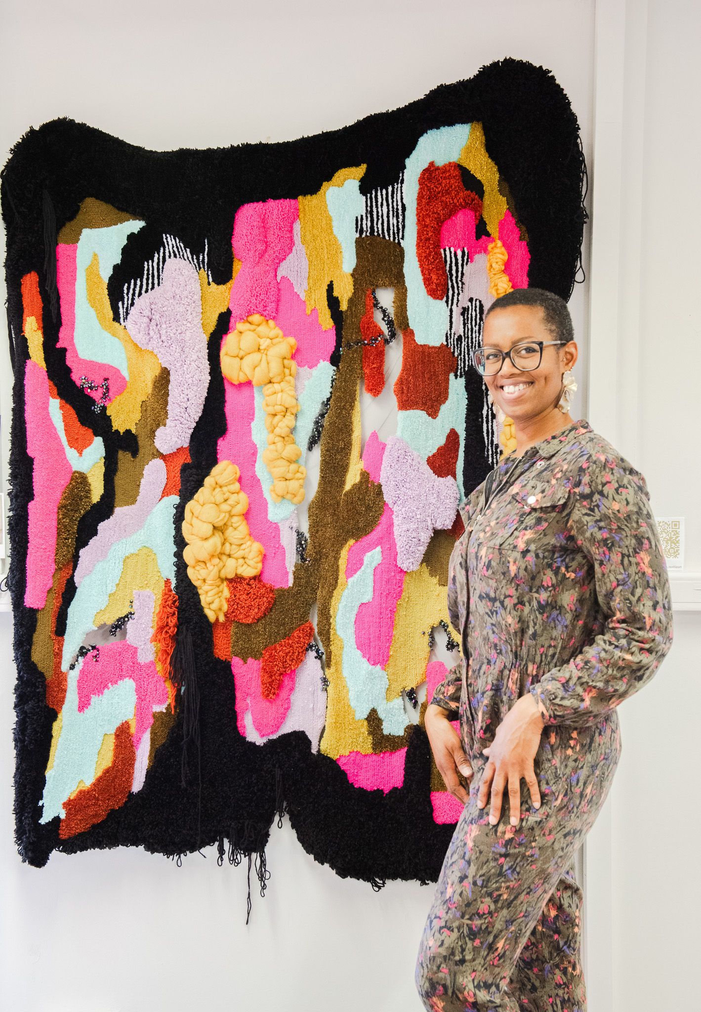 Artist posing beside large colorful abstract textile wall hanging with mixed yarn textures.