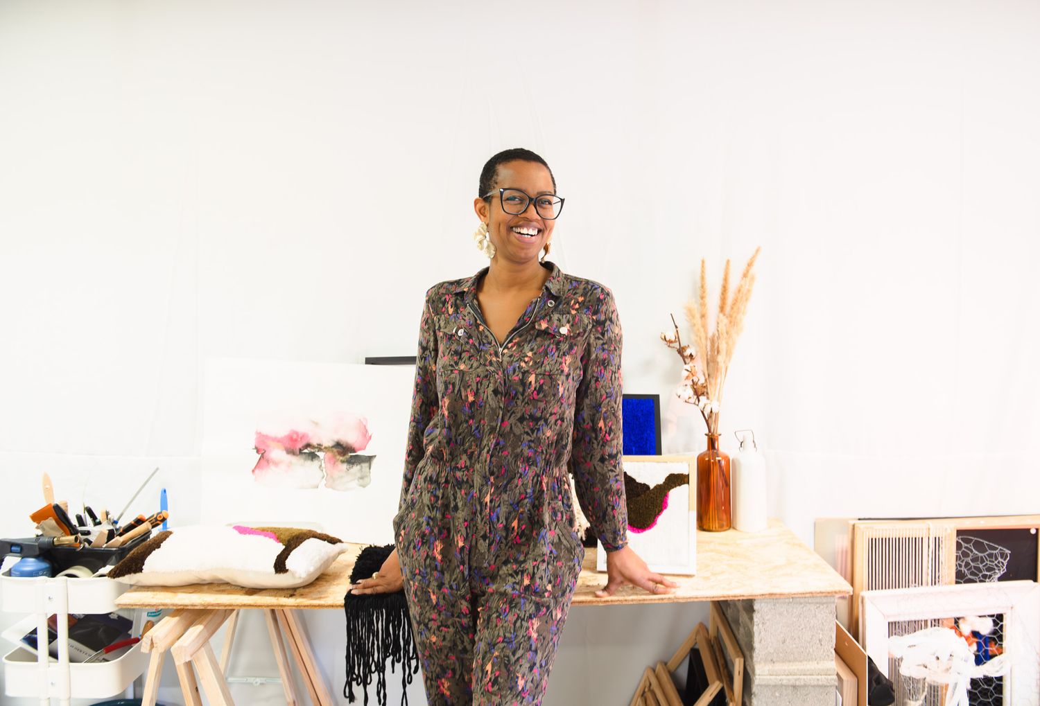 Woman in floral jumpsuit smiling in bright art studio workspace with wooden tables and decorative items.