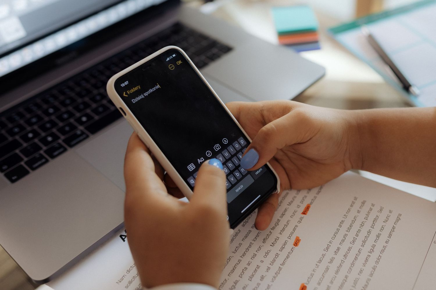 Person typing on smartphone near laptop and documents on desk.