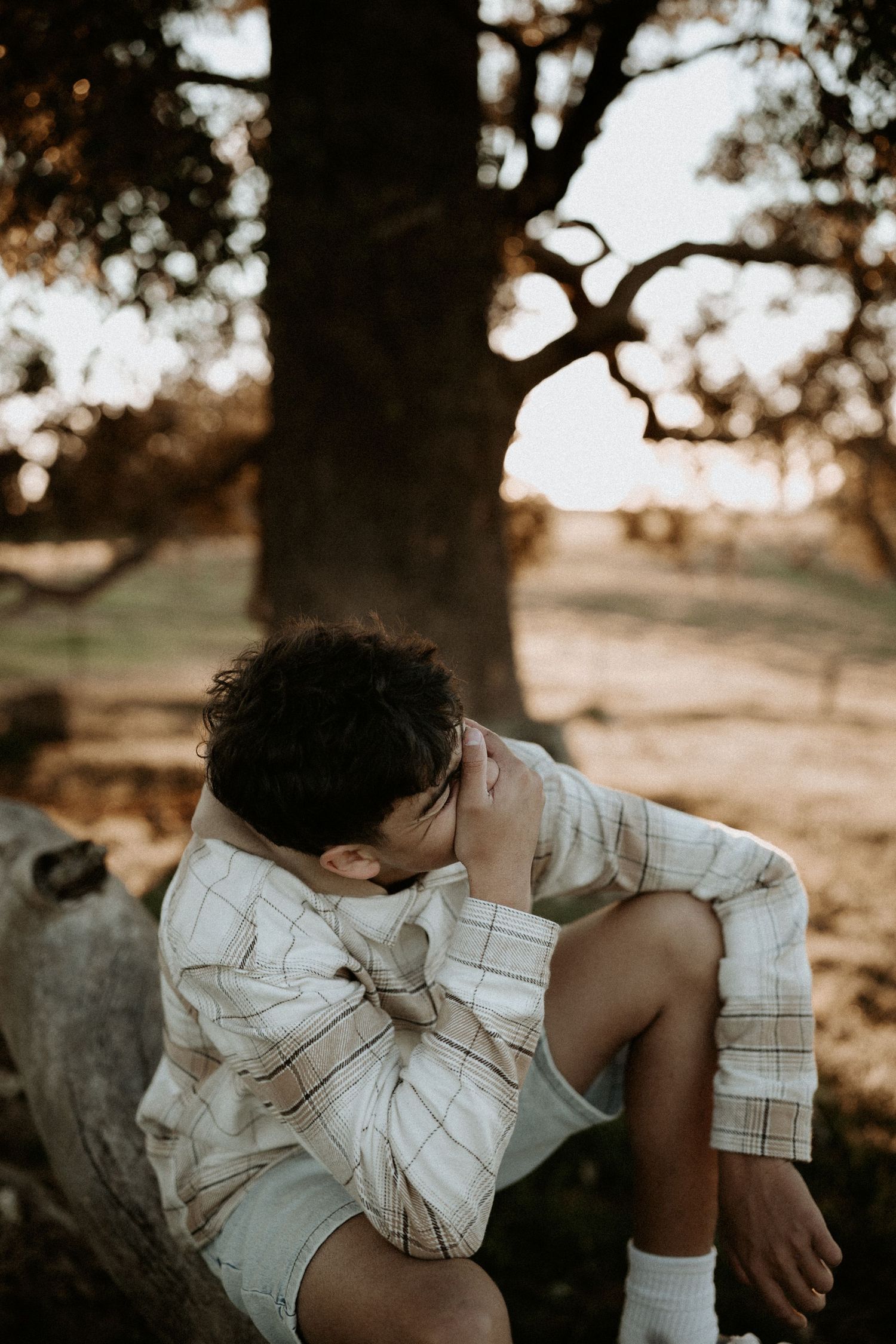 Young man sitting on a log under a tree at sunset, wearing a plaid shirt, with his face resting in his hand whilst laughing.