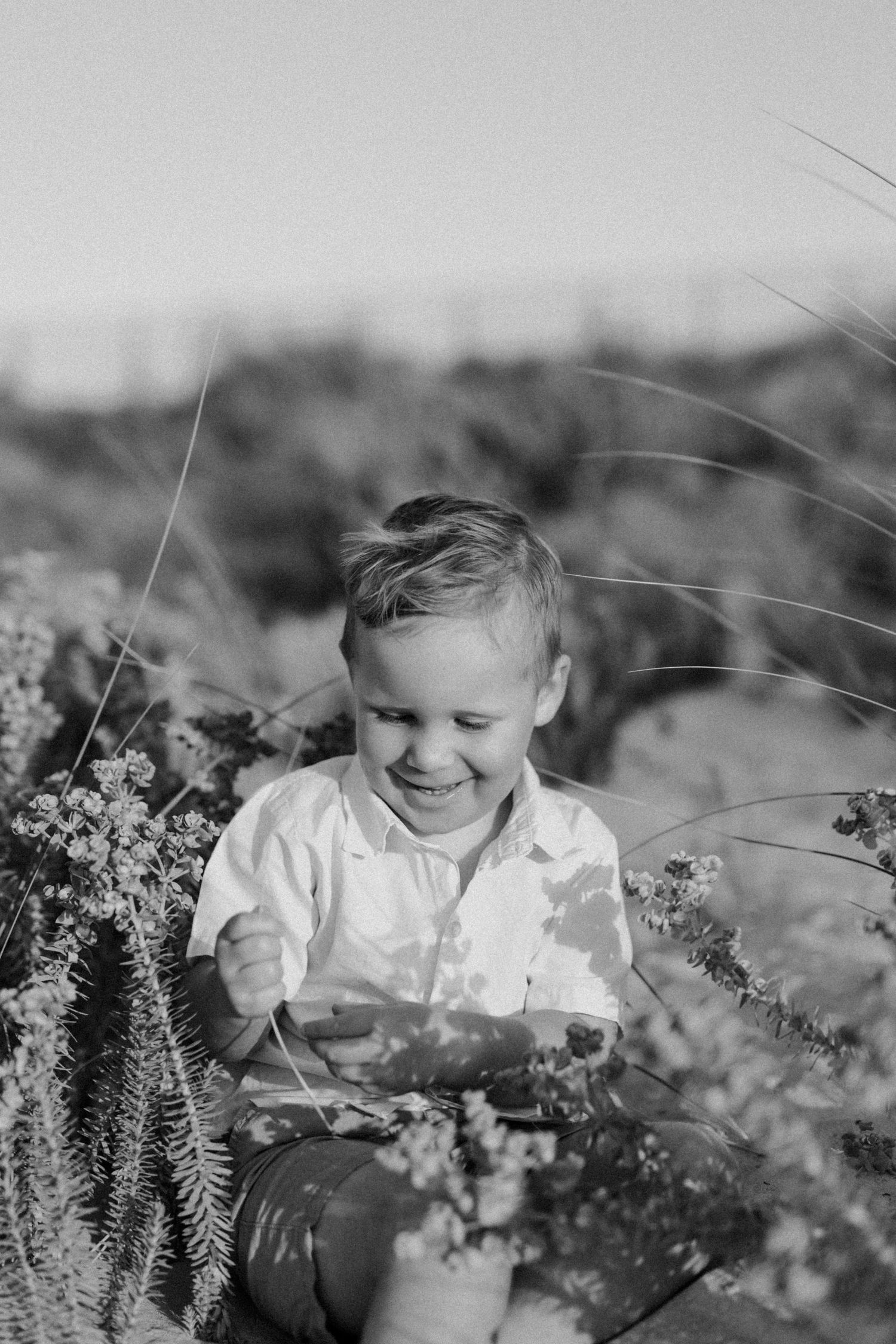 Black and white photo of a happy young boy smiling while sitting at the beach during his family session in Bunbury, Western Australia.
