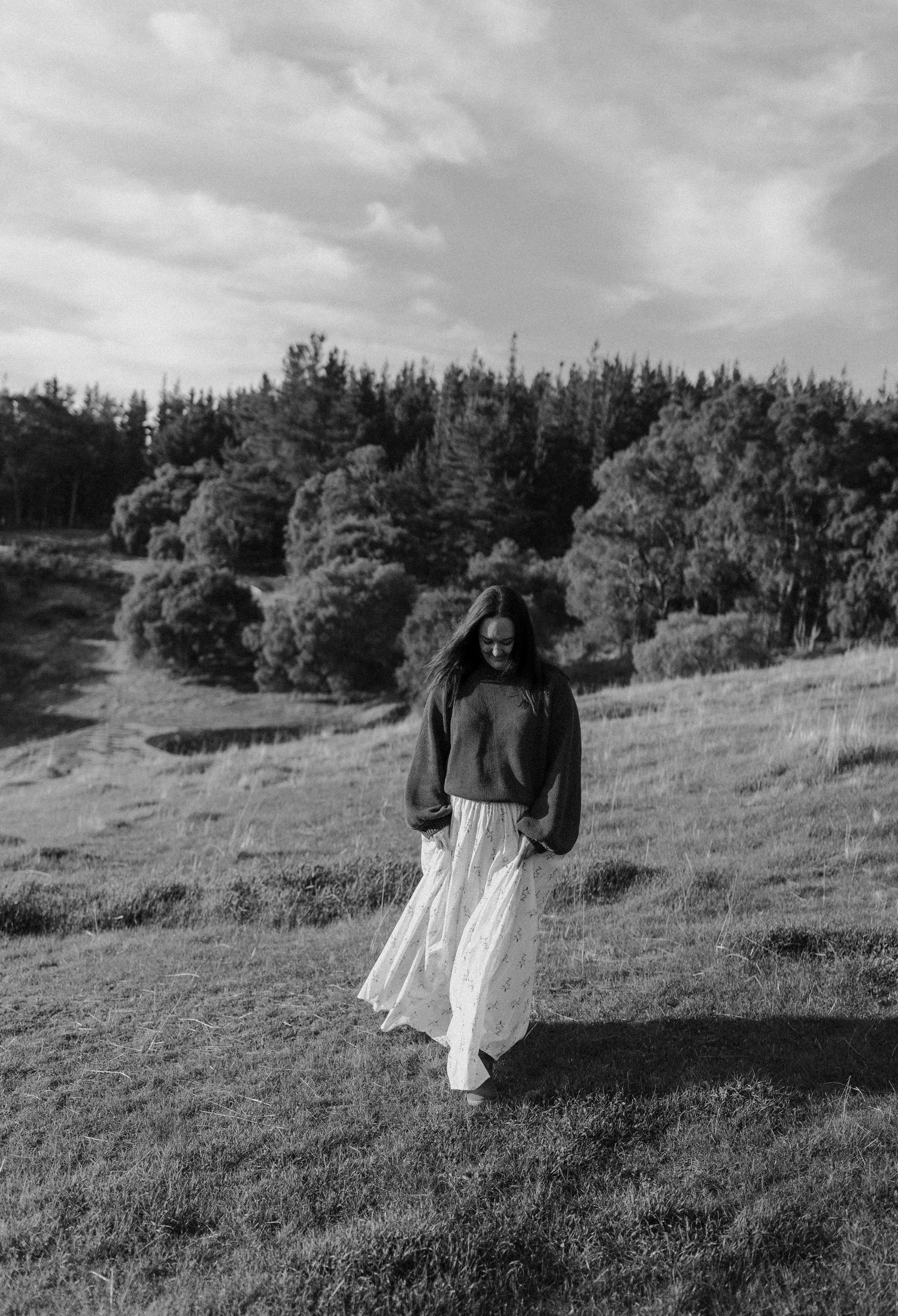 Black and white photo of a woman in a flowing white skirt and dark sweater walking through a lush green meadow.