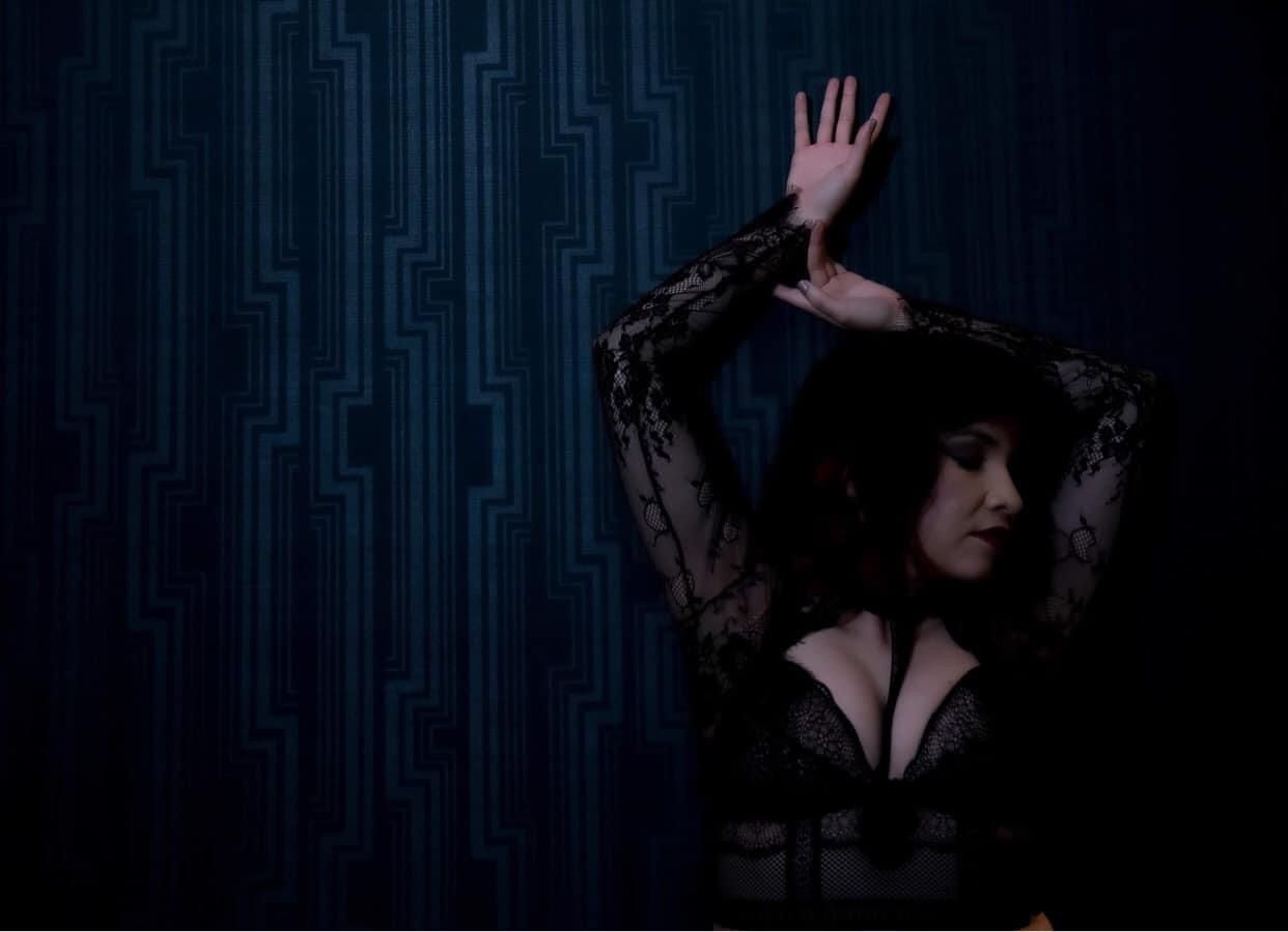 Moody image of a Latina woman hands above her head and lacy lingerie set on a dark blue wall. Boudoir Portrait Session by Hinton Imagery