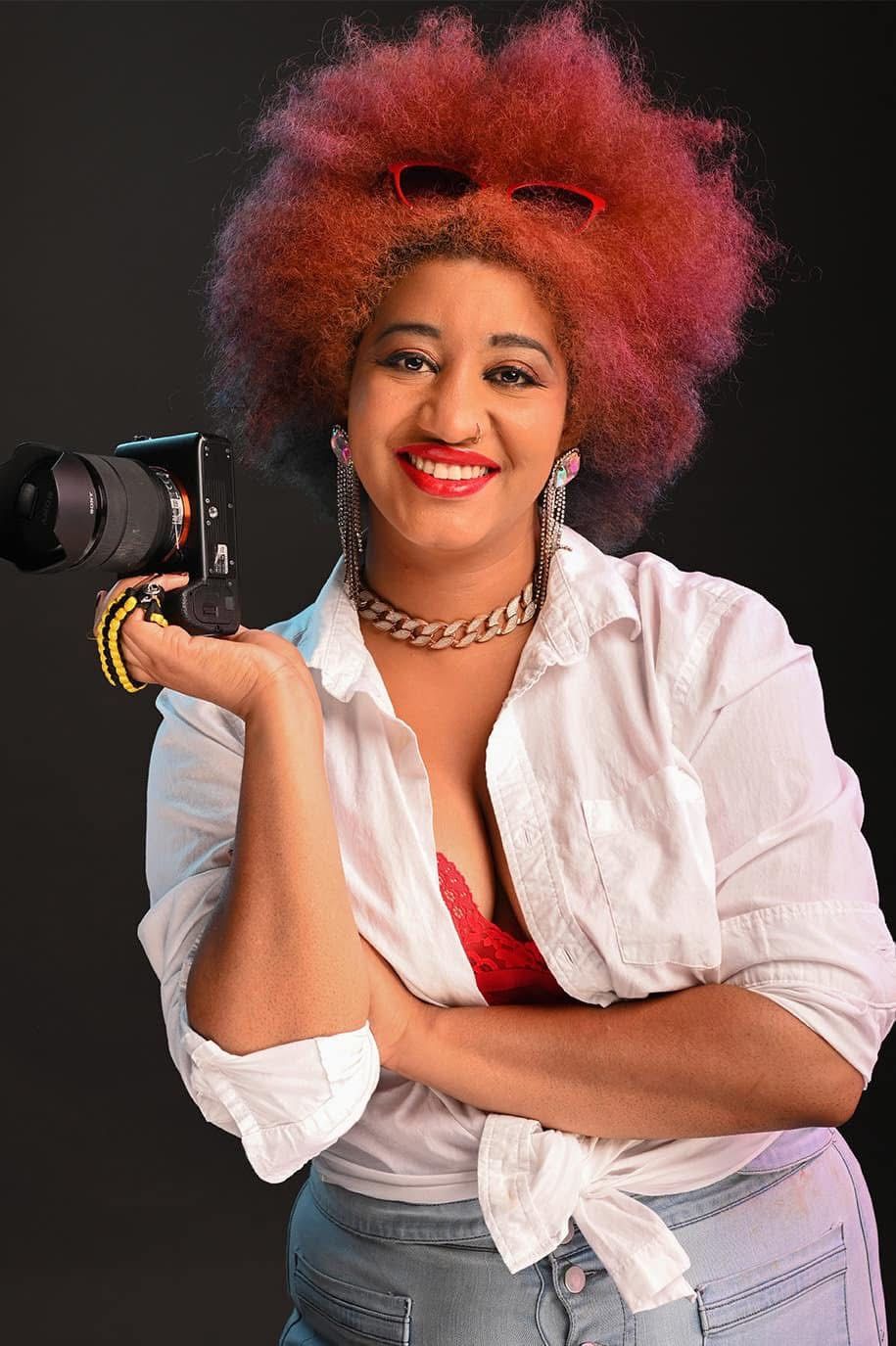 Image of Hinton the photographer behind Hinton imagery, she has a huge red afro here and is smiling while holding a camera.