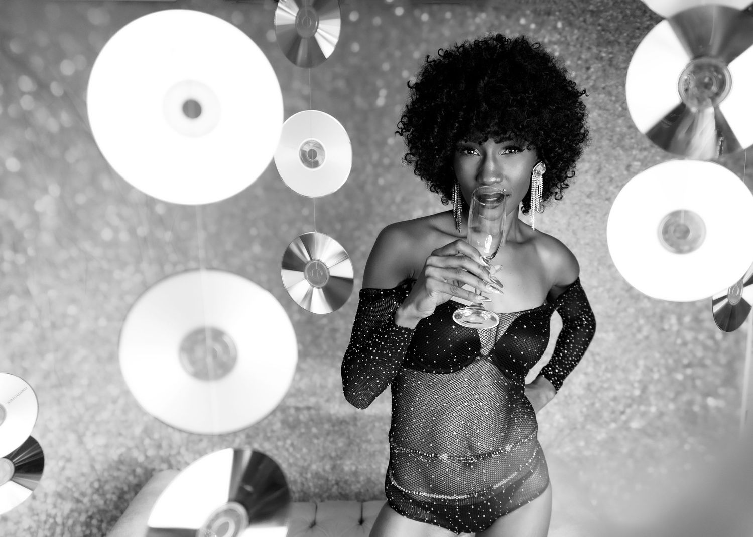 Black & white photo of Boudoir session in Jacksonville NC. Black woman with an Afro holding a glass in a sparkly bodysuit up to her lips.