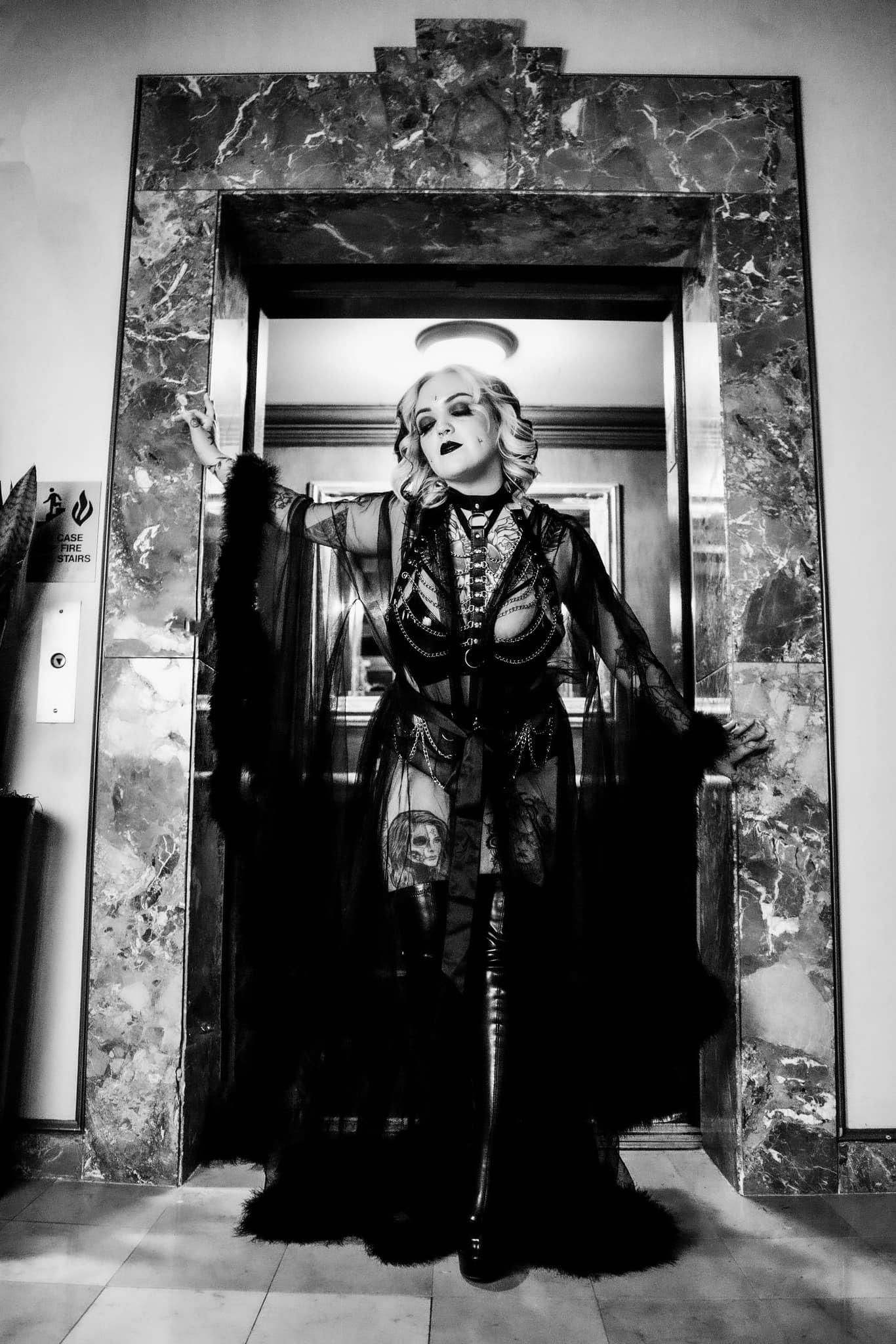 Black and White image of a Heavily tattooed White woman in a luxury black robe stepping forward as light silhouettes her. Boudoir by Hinton
