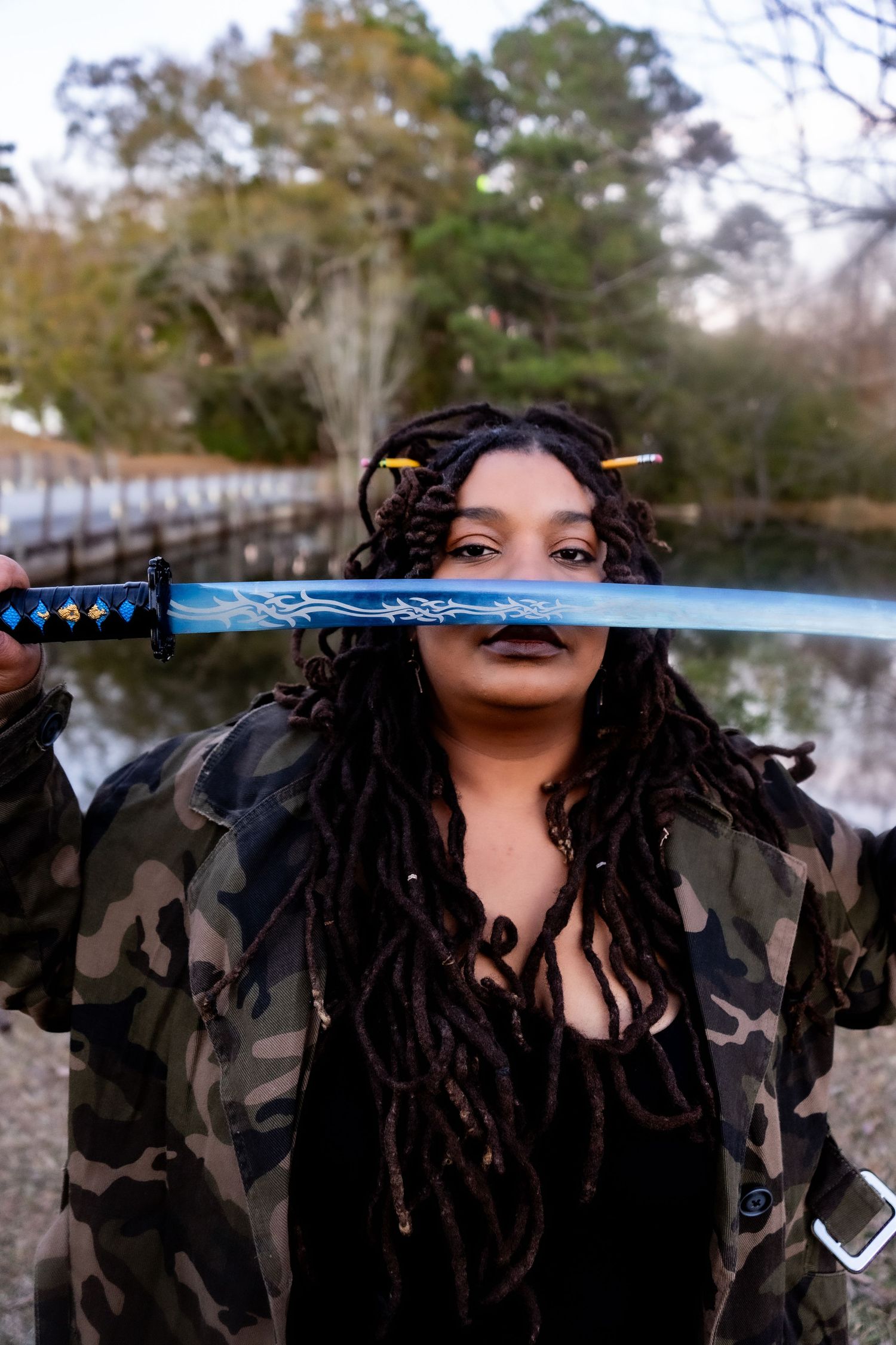 Photo of a gorgeous Black woman with locs and pencils in her hair. She is outside with a samurai sword in front of her face. Stern look