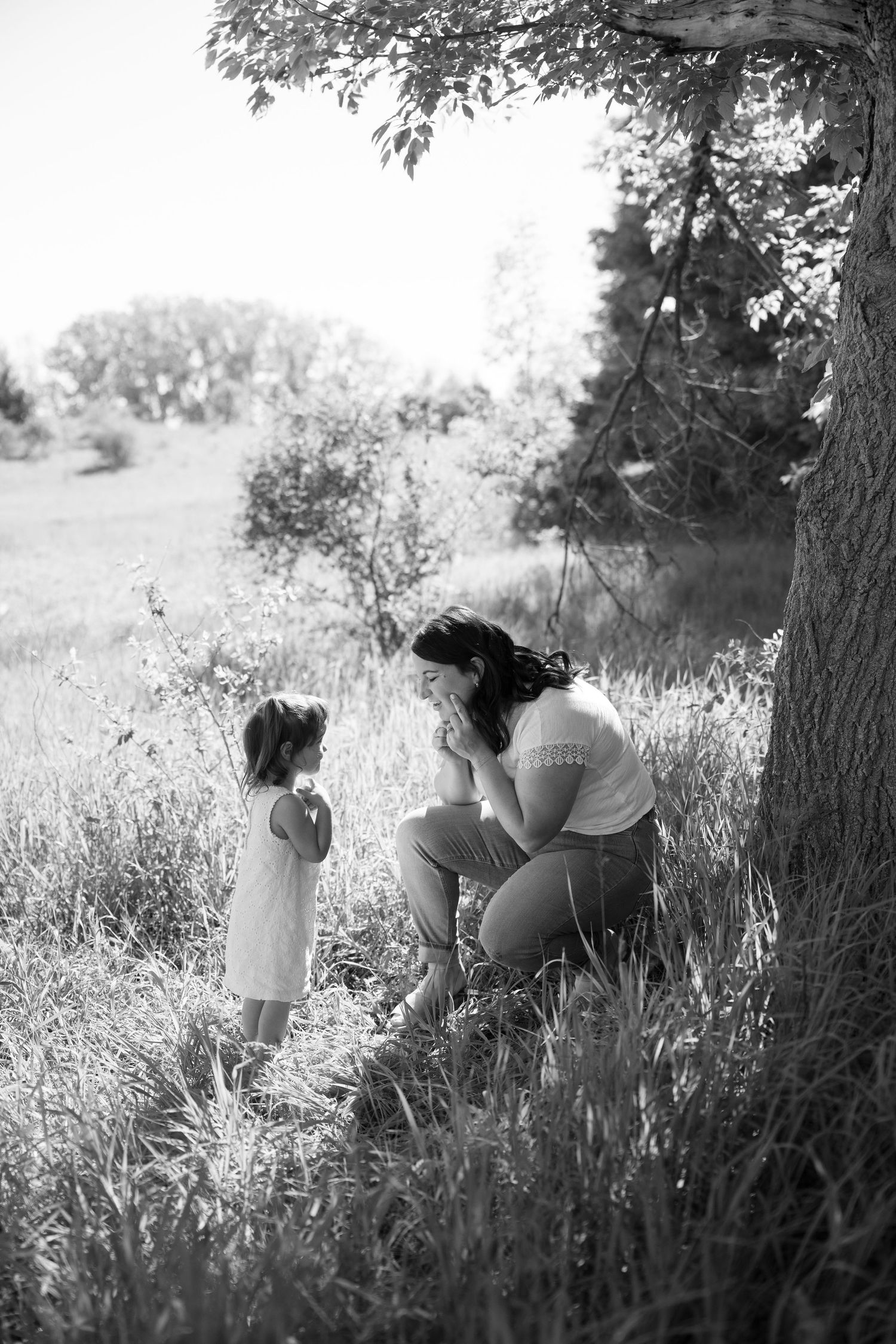 Black and white photo of a mother crouching beside a young girl near a tree in a sunlit meadow.