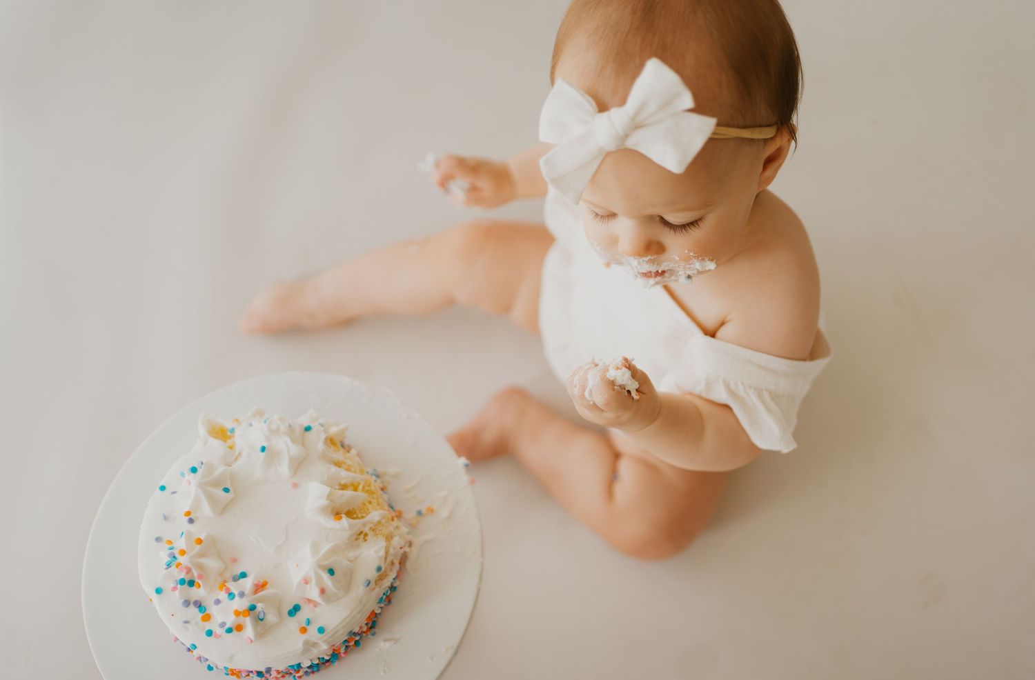 Baby in white outfit with bow sits next to decorated birthday cake.