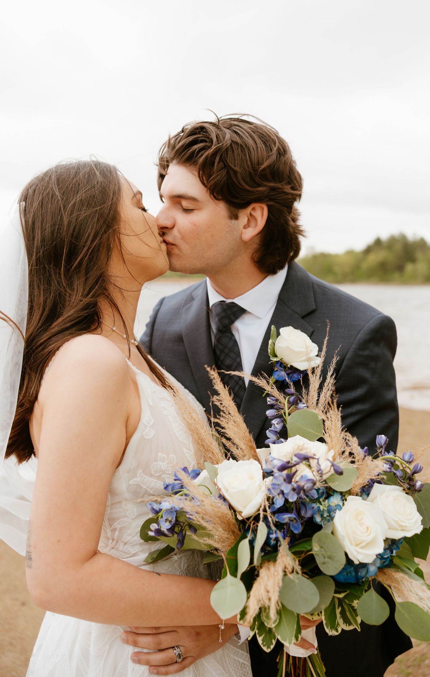 Romantic wedding couple shares intimate kisses with a beautiful bouquet of white roses and blue flowers.