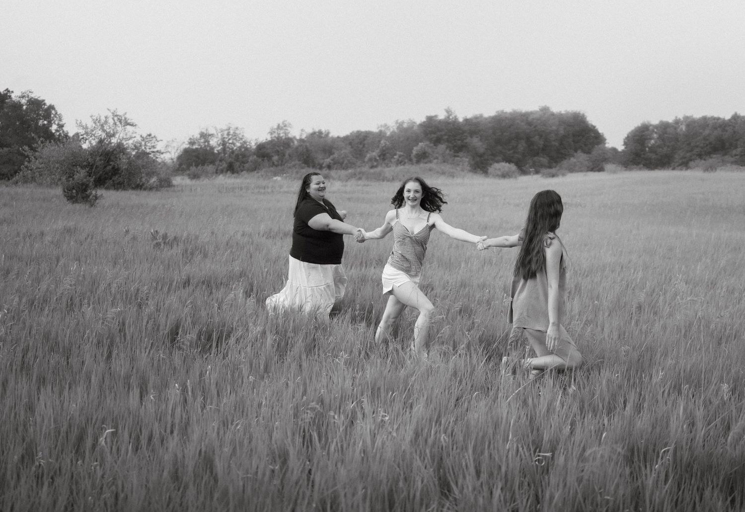 Three girls dancing joyfully in a grassy meadow, black and white photo capturing carefree summer fun outdoors.