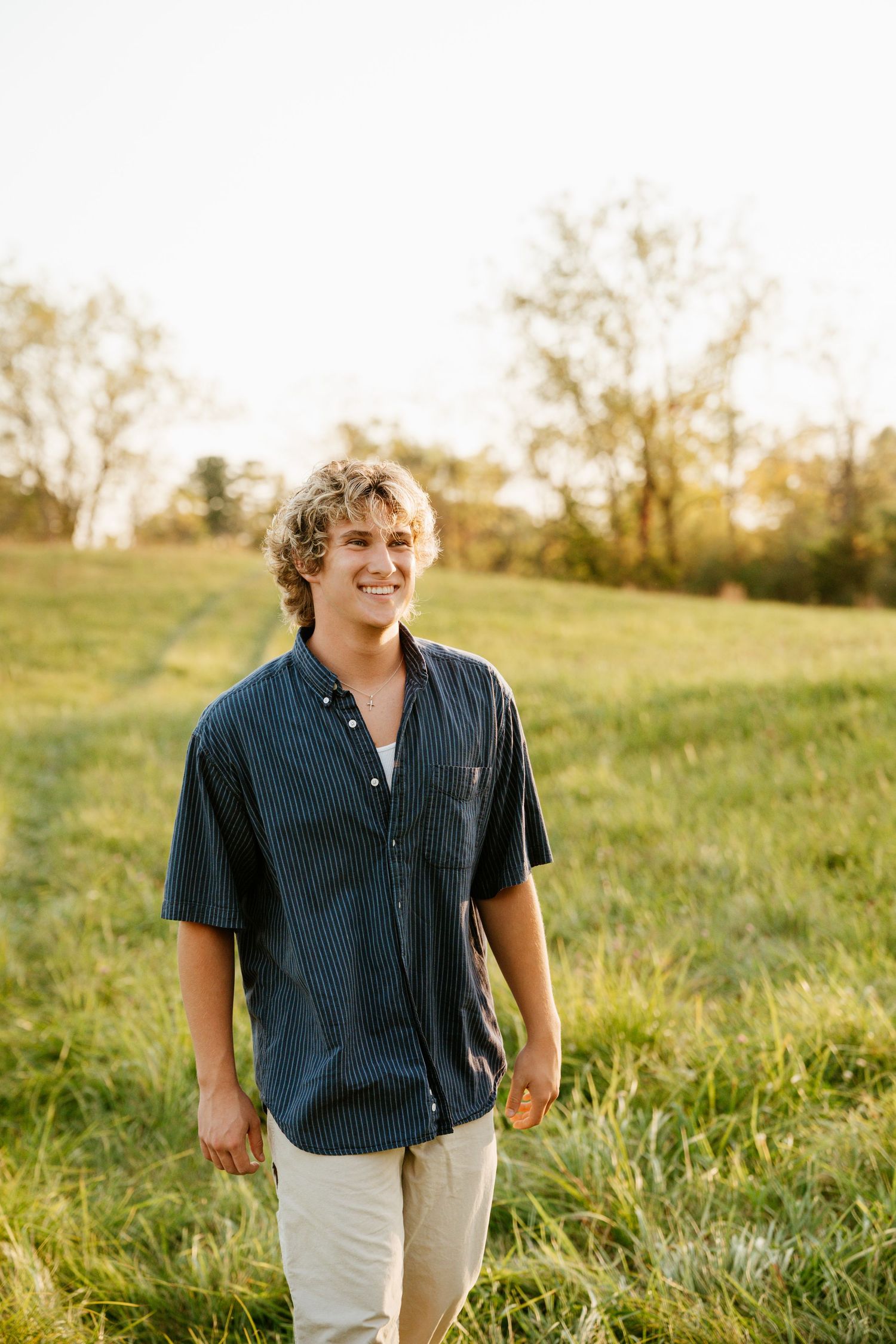 Young man with curly blond hair smiling in a green field, wearing a navy short-sleeve shirt and khaki pants.
