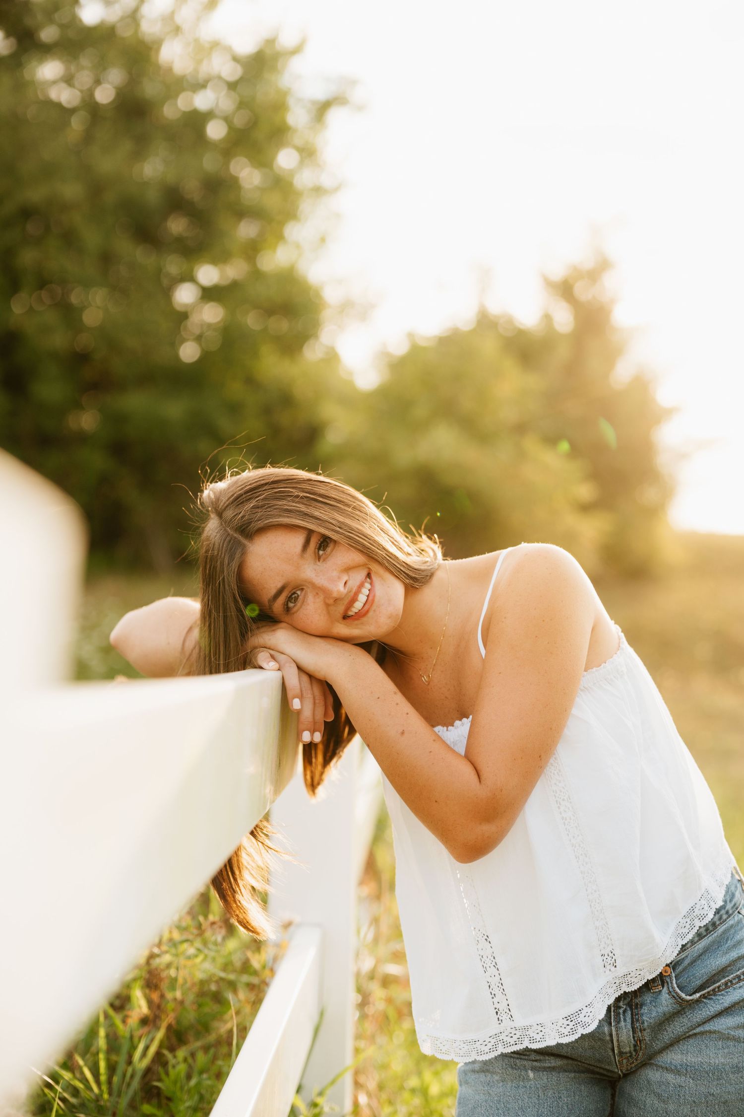 Young woman in white top and jeans leaning on a fence, smiling outdoors in golden sunlight.