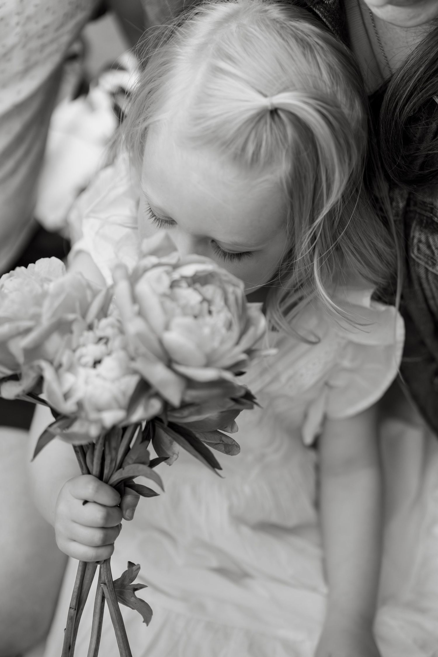 Black and white photo of a young flower girl smelling a bouquet of peonies at a wedding.