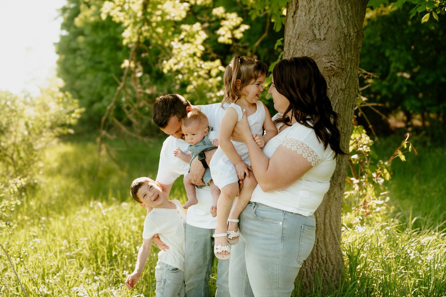 Family of five posing by a tree in a lush green park, wearing white tops and light jeans.