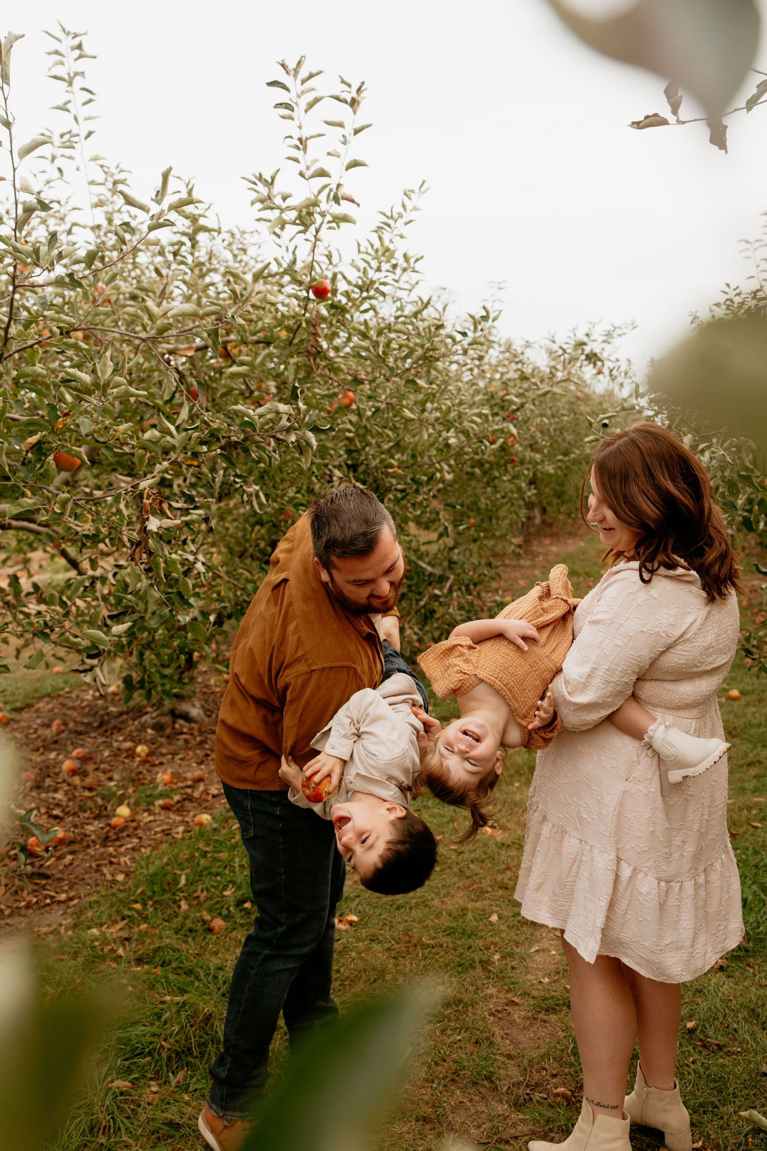 Family playfully swinging child upside down in an apple orchard during a fall outdoor photo session.