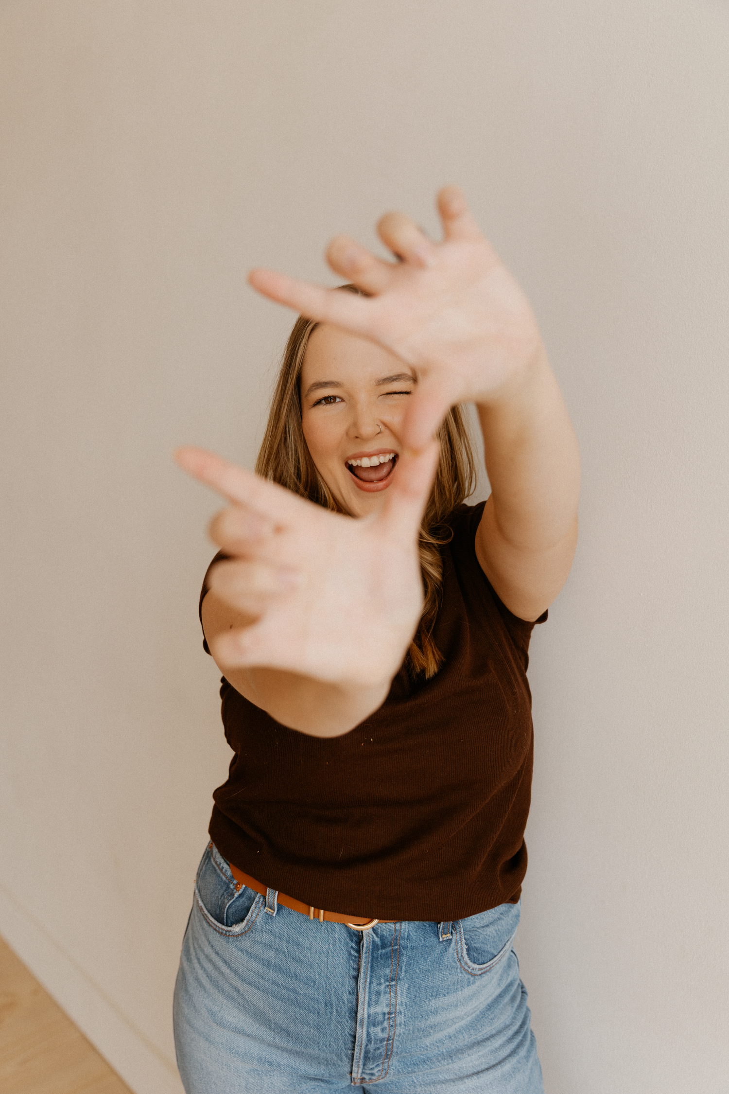 Person wearing black t-shirt and jeans making playful hand gestures against beige wall.