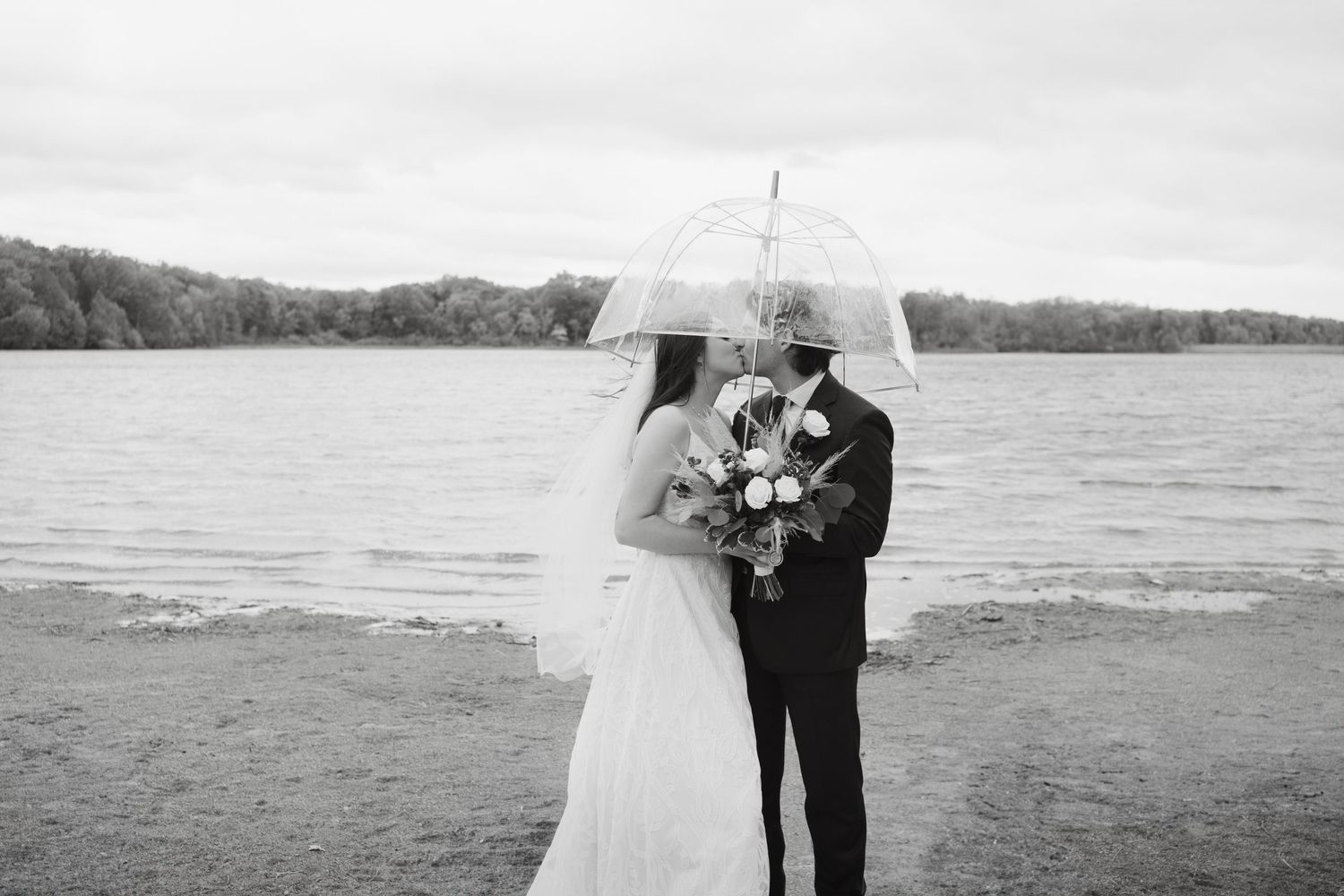 A couple shares an intimate moment under white umbrellas on a cloudy beach during their wedding photoshoot.