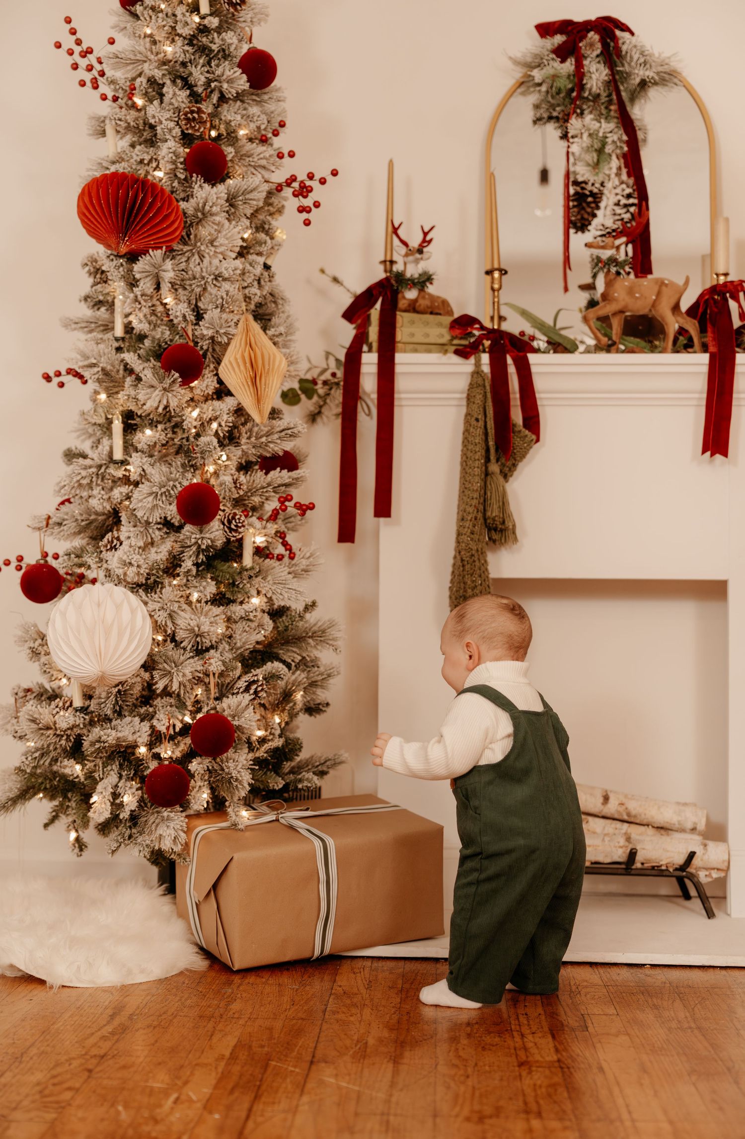 A baby in green overalls stands by a flocked Christmas tree with red ornaments next to a decorated fireplace mantel.