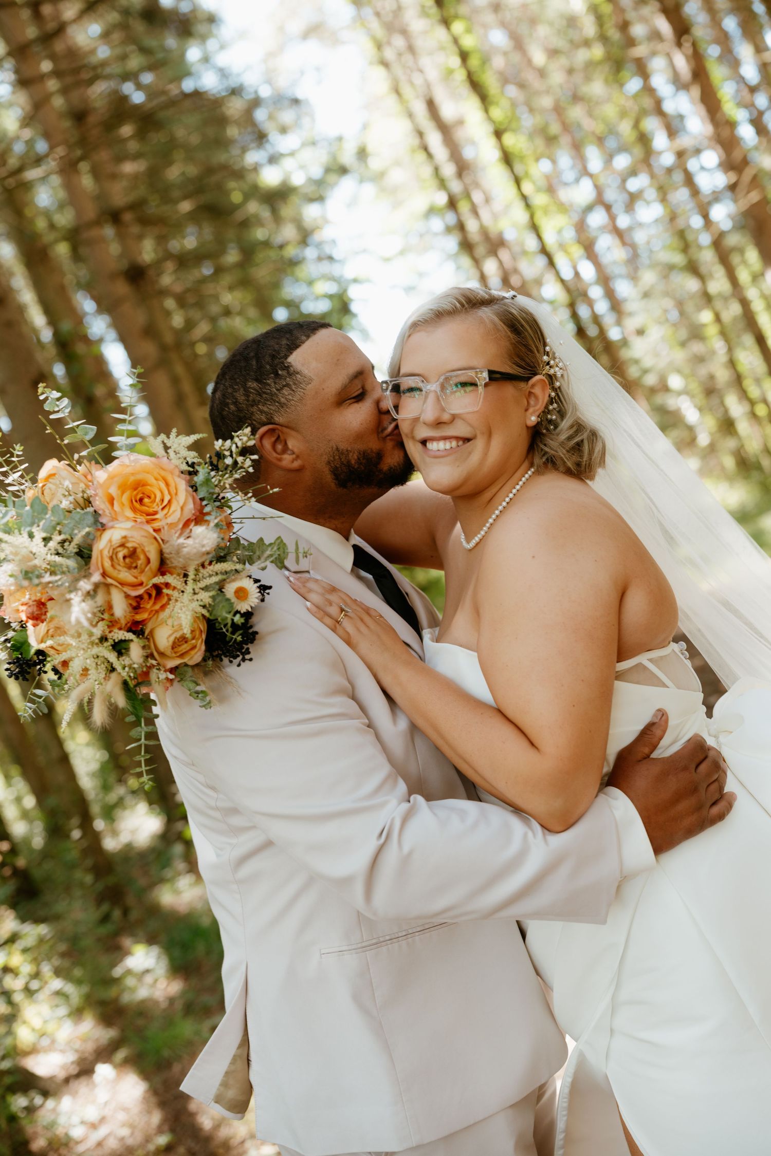 A groom in a white suit kisses his smiling bride holding an orange floral bouquet in a sunlit forest setting.