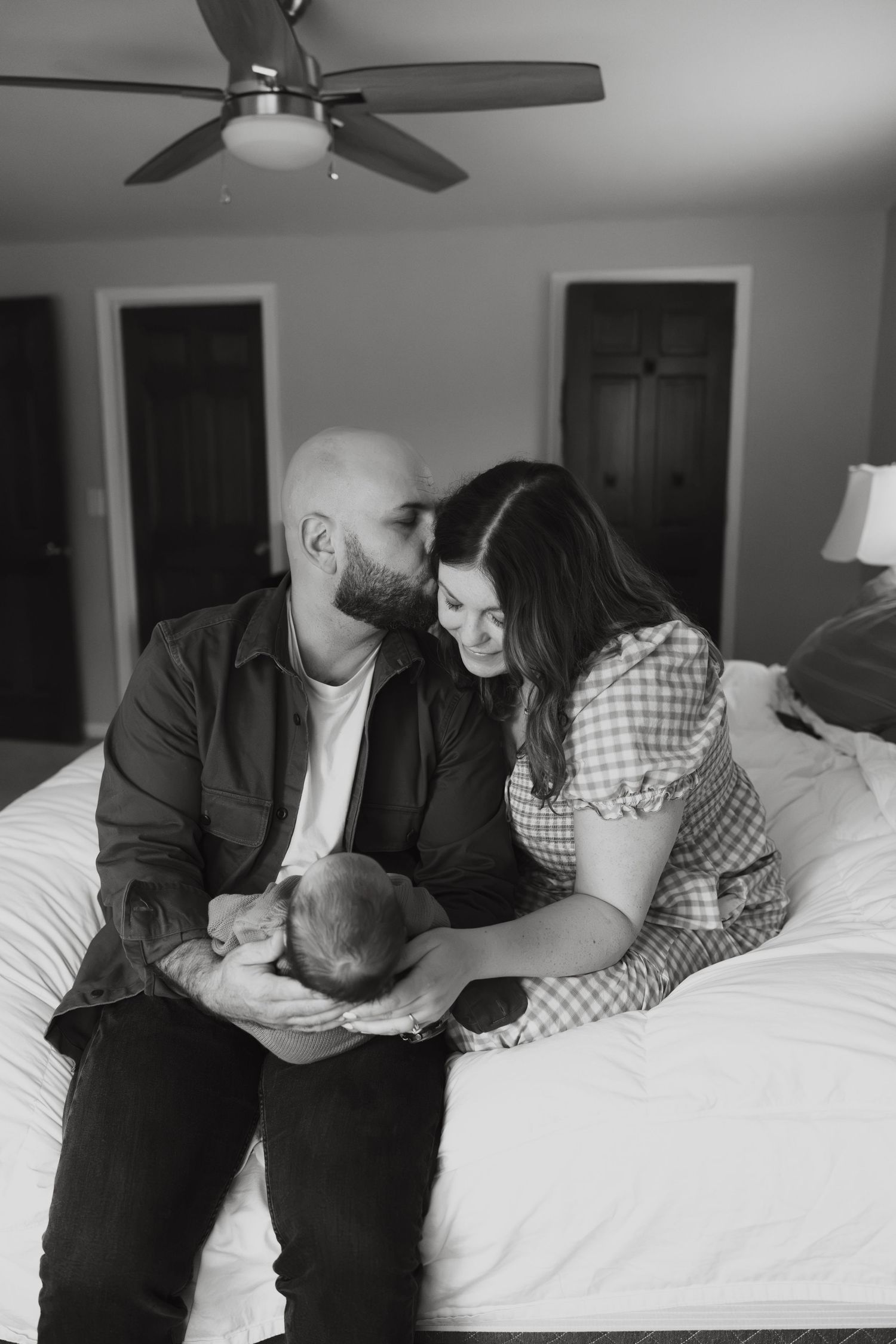 Black and white photo of a couple sitting on a bed, sharing a tender moment while cradling their newborn baby.