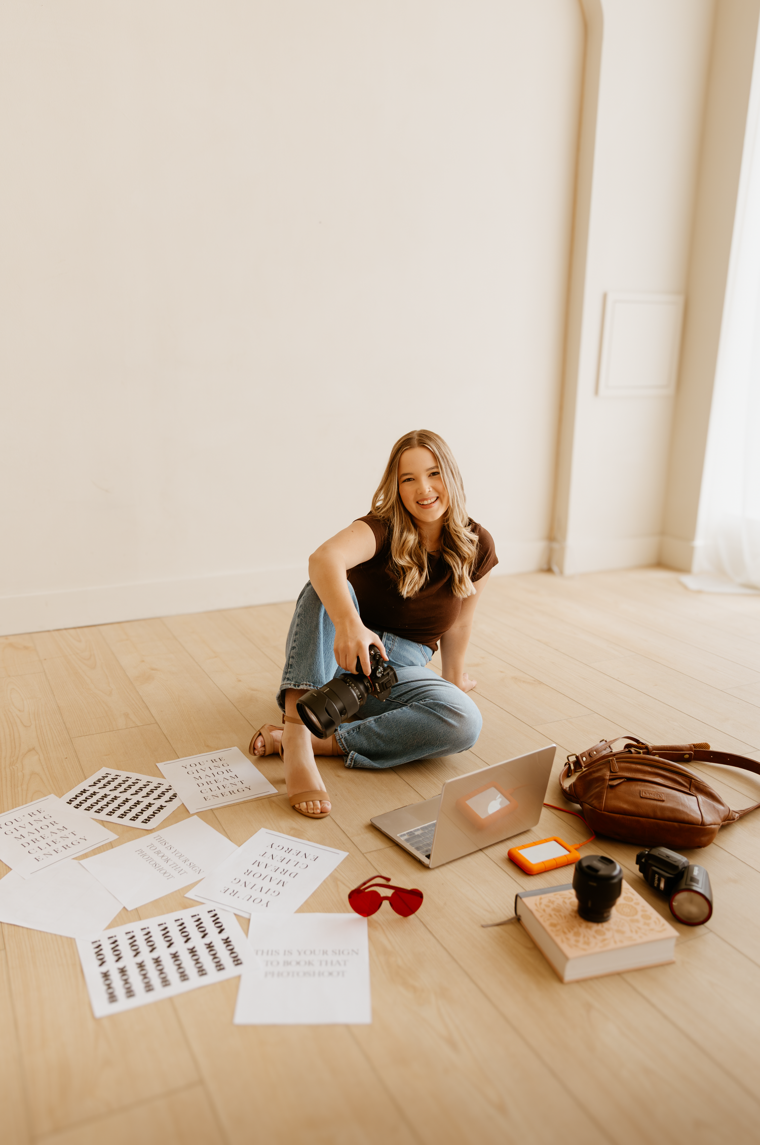 Person sitting on wooden floor with laptop, papers, and brown leather bag in minimalist room.
