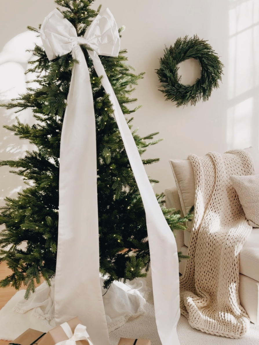 A decorated Christmas tree with a large white bow, green wreath on wall, and chunky knit blanket on a cream sofa.