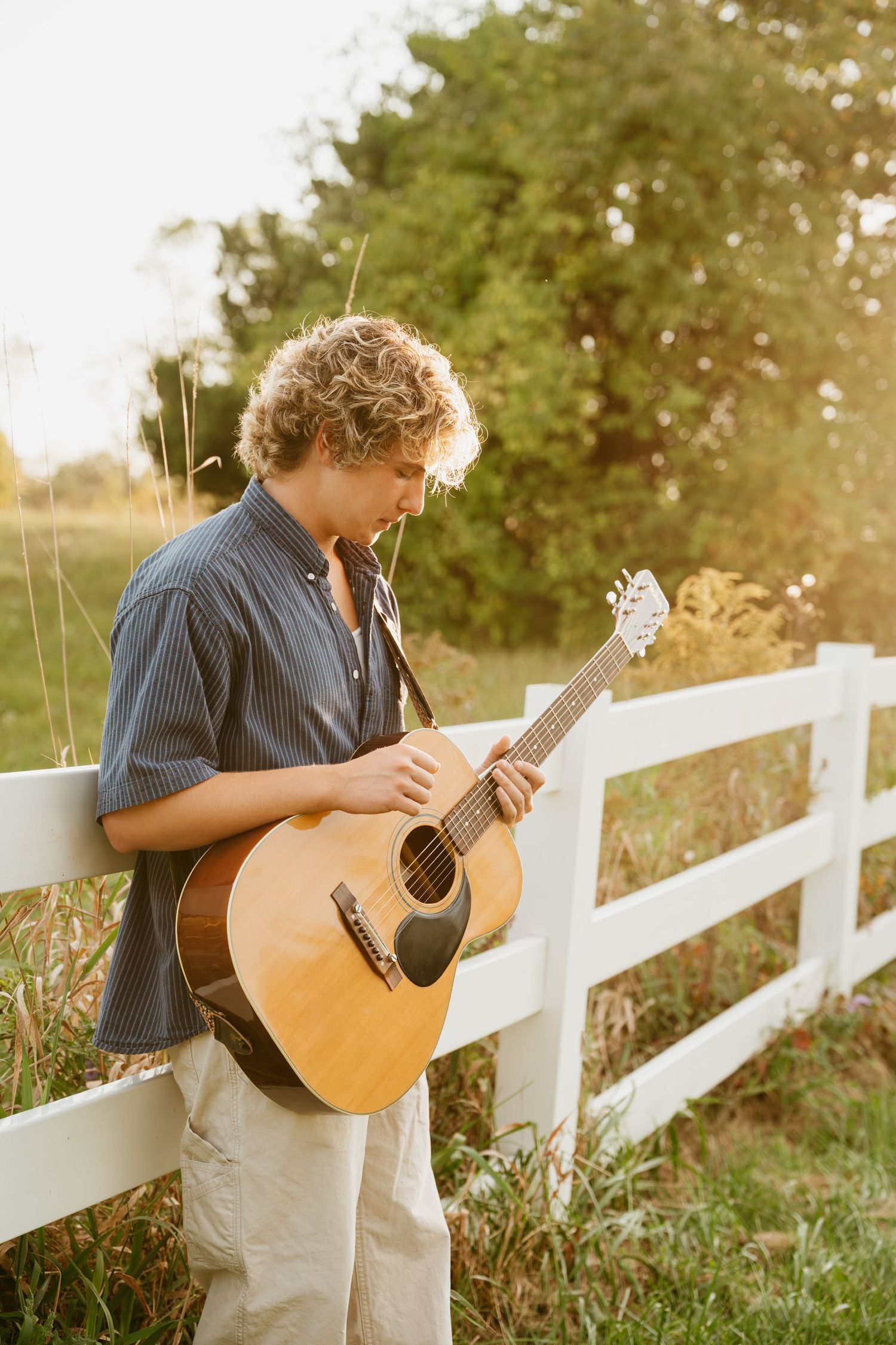 Person plays acoustic guitar by white fence during golden sunset.