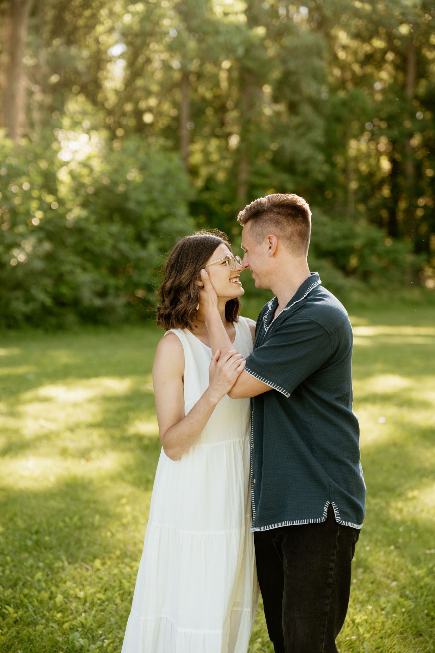 A couple shares a romantic moment in a sunlit green park, the woman in a white dress smiling up at a man in a dark shirt.